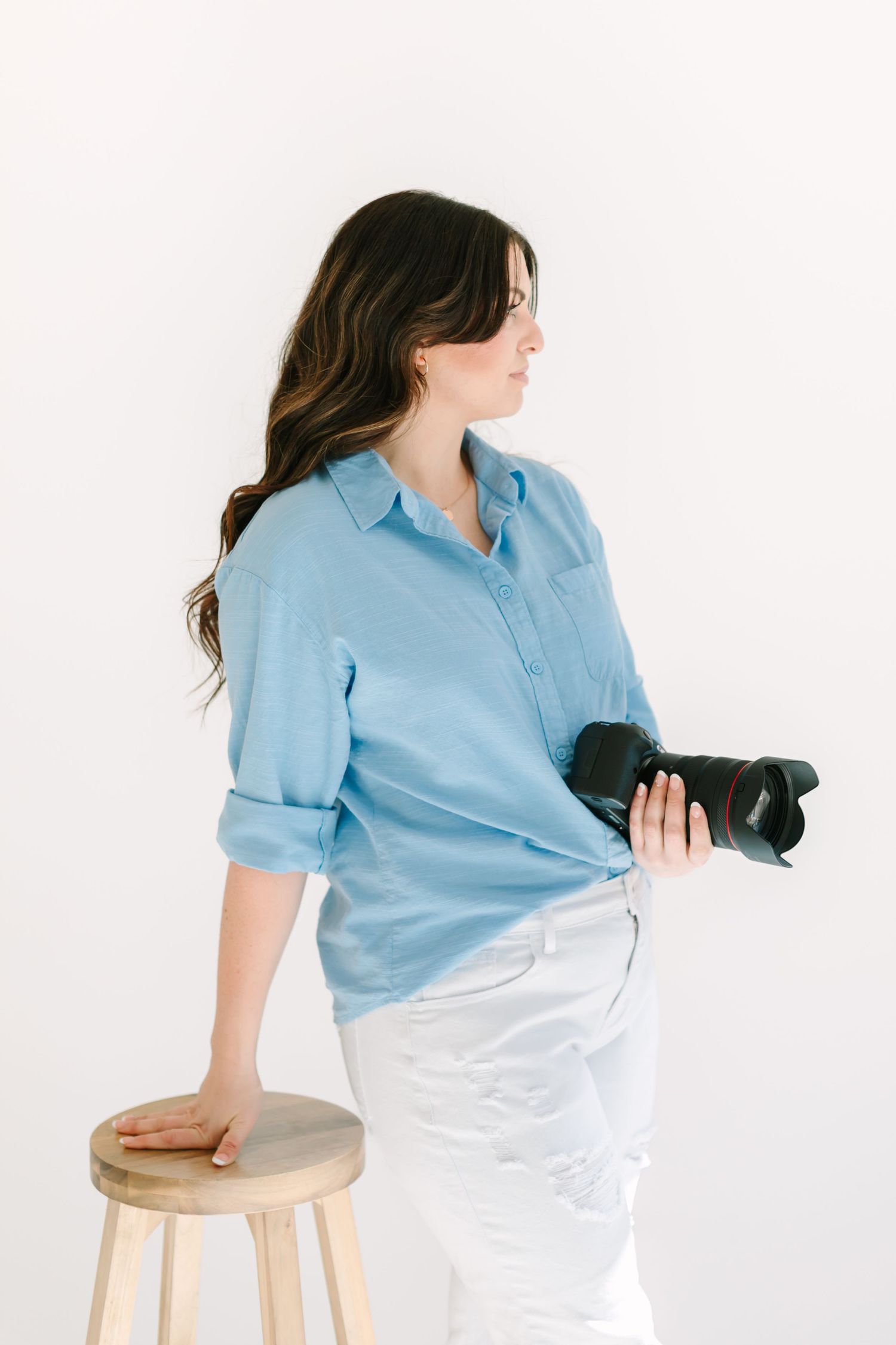 Woman in light blue shirt and white pants sits casually on wooden stool in minimalist studio setting.