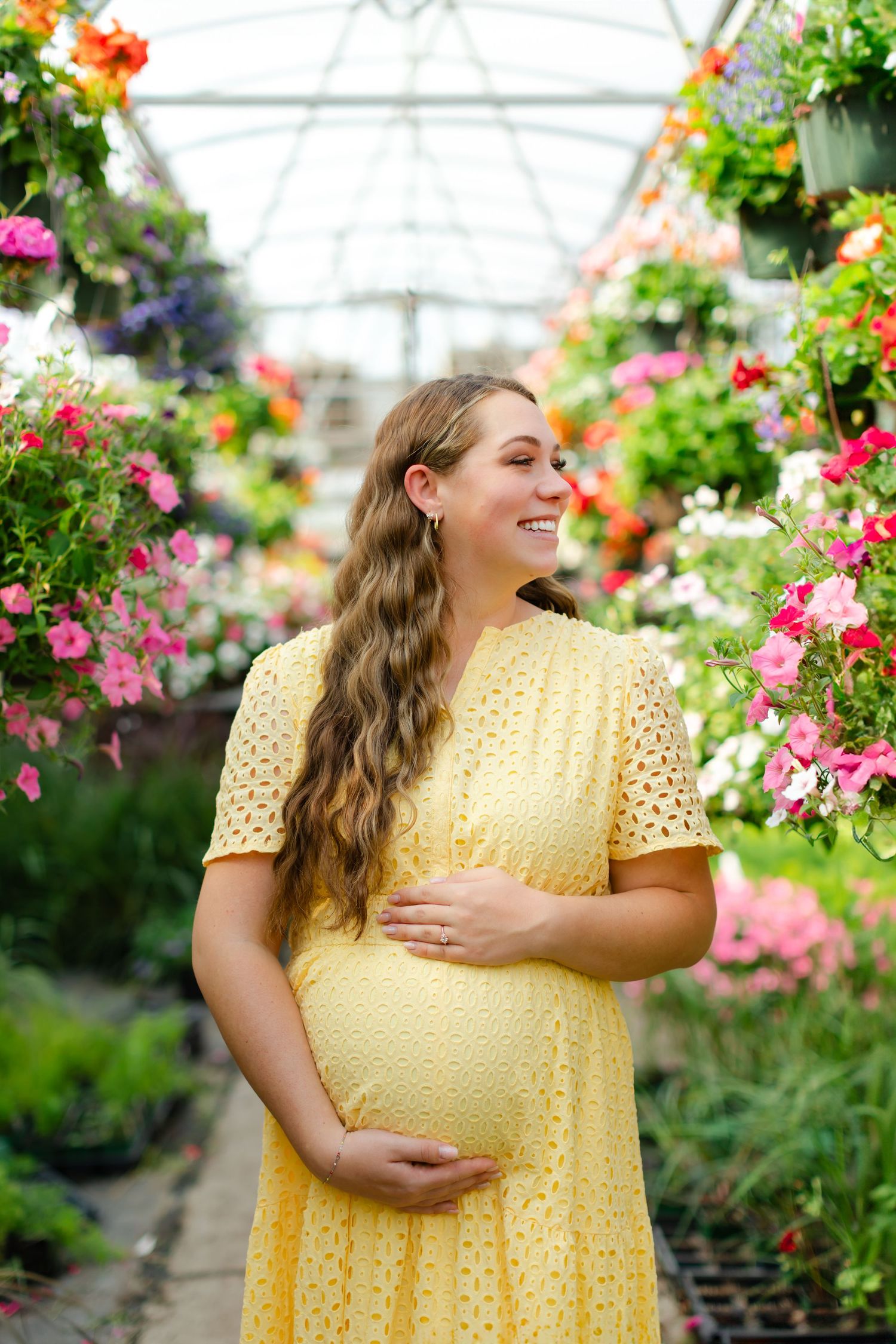 A person in a yellow polka dot dress poses among colorful hanging flower baskets in a greenhouse.