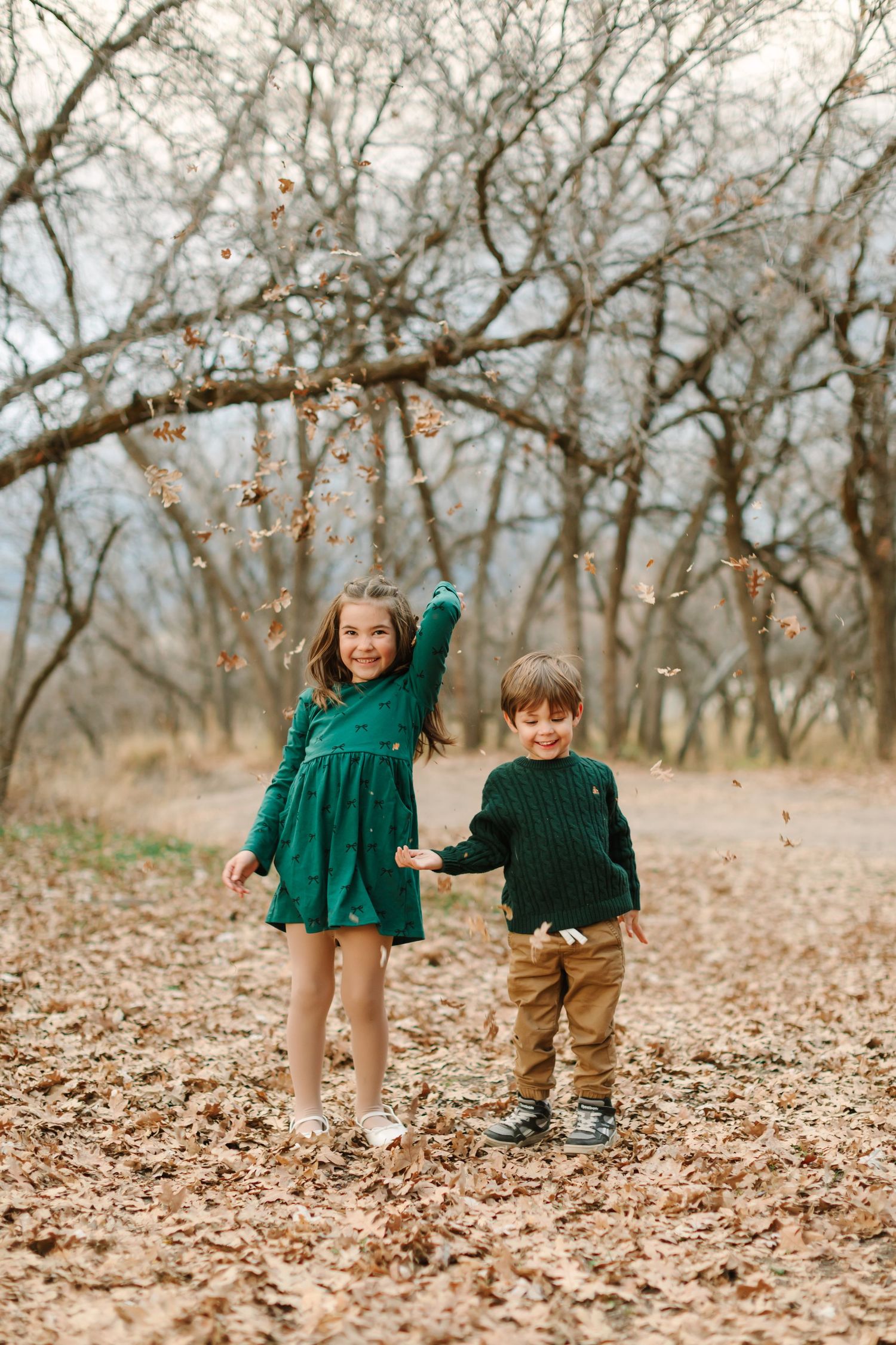 Two children in green and brown outfits playing together in autumn leaves on a forest floor.
