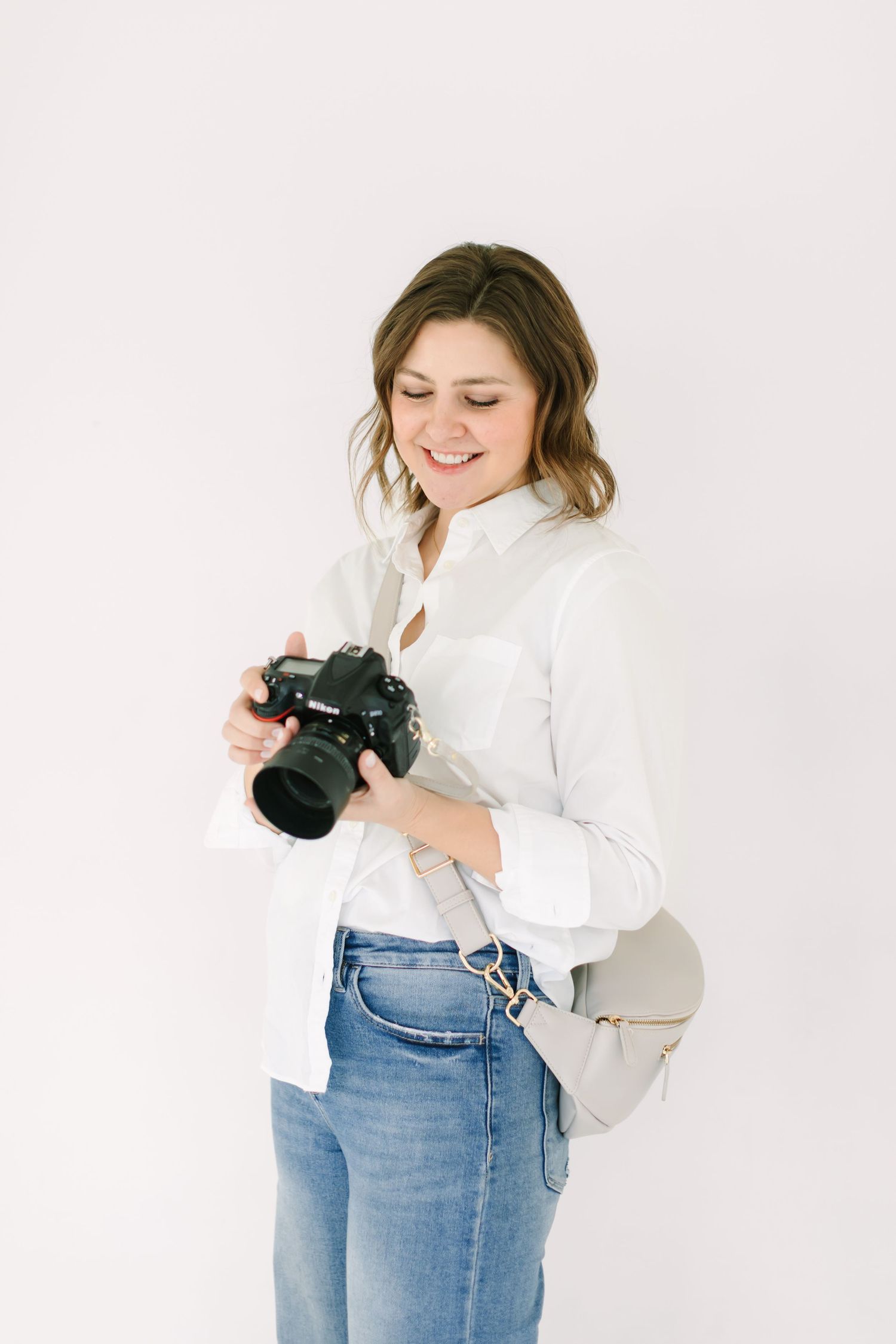 Professional photographer in white sweater and jeans holds camera while smiling and posing against white backdrop.