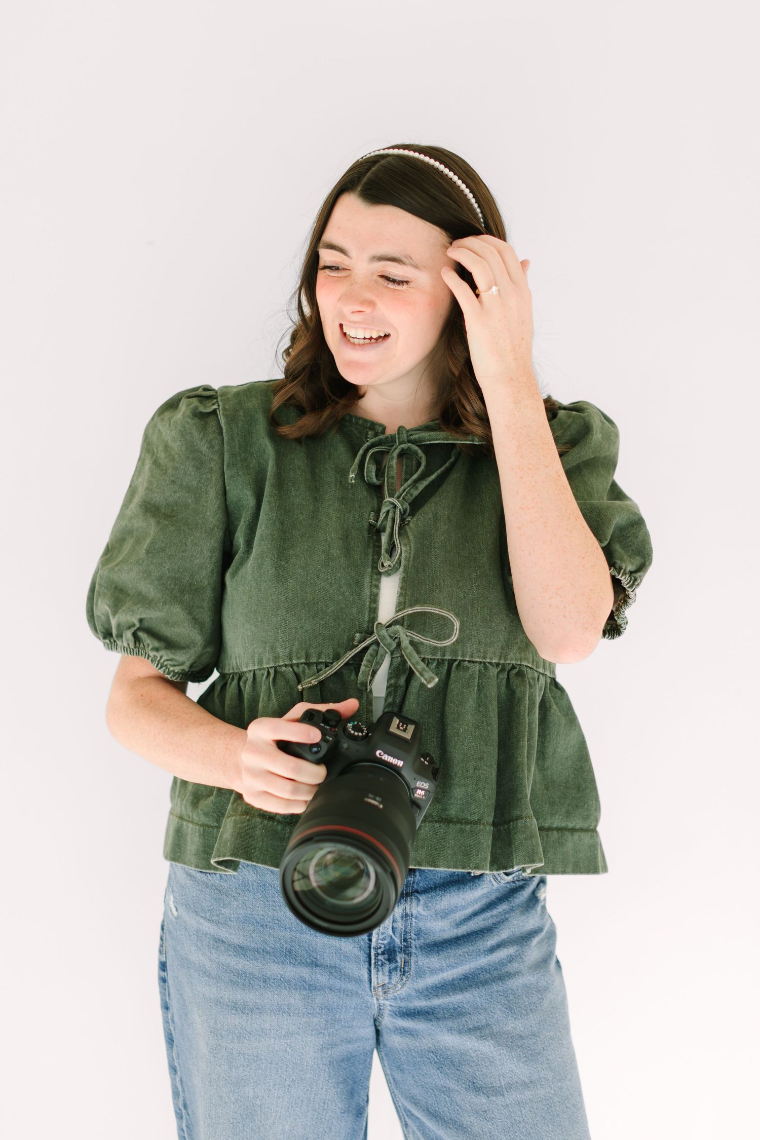 Photographer in green puff sleeve top and jeans laughs while holding professional camera against white background.