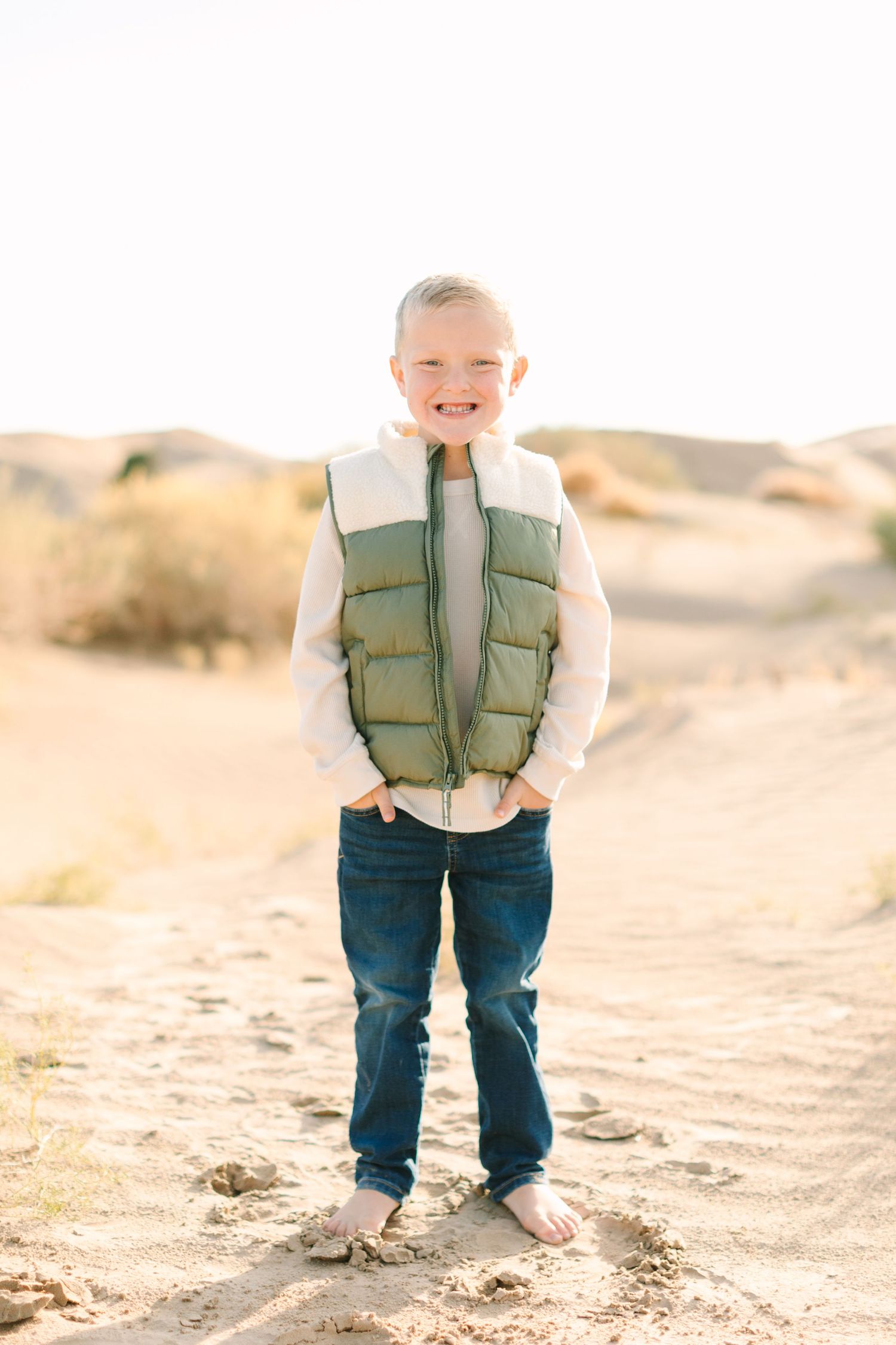 A child wearing a green puffer vest and jeans stands smiling in a desert landscape on a sunny day.