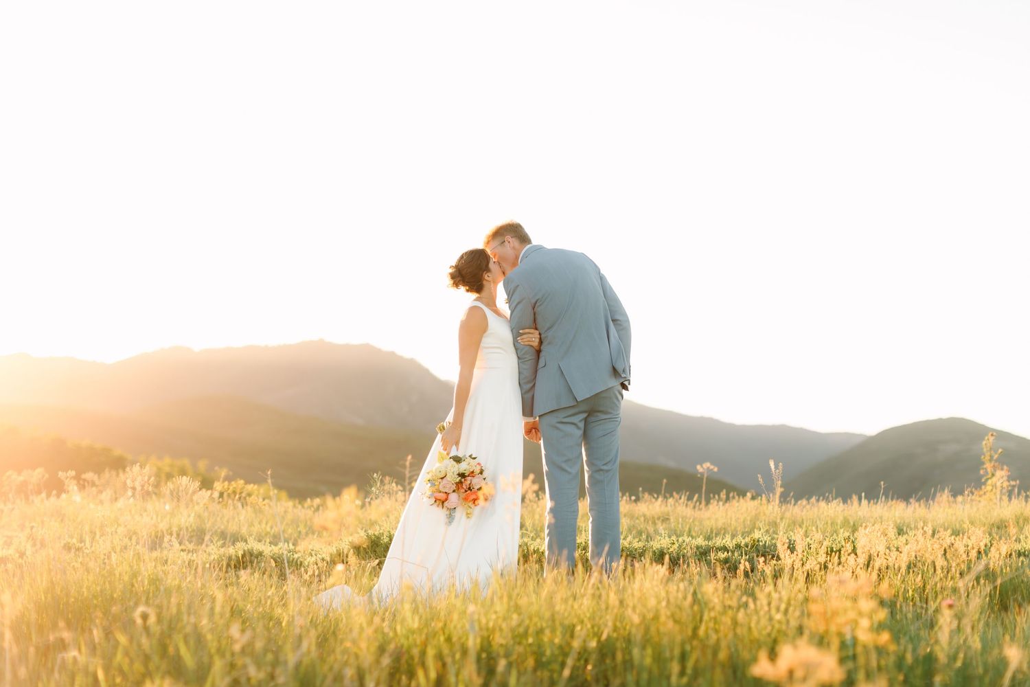 Bride and groom kissing in golden sunlit meadow with mountain backdrop on their wedding day.