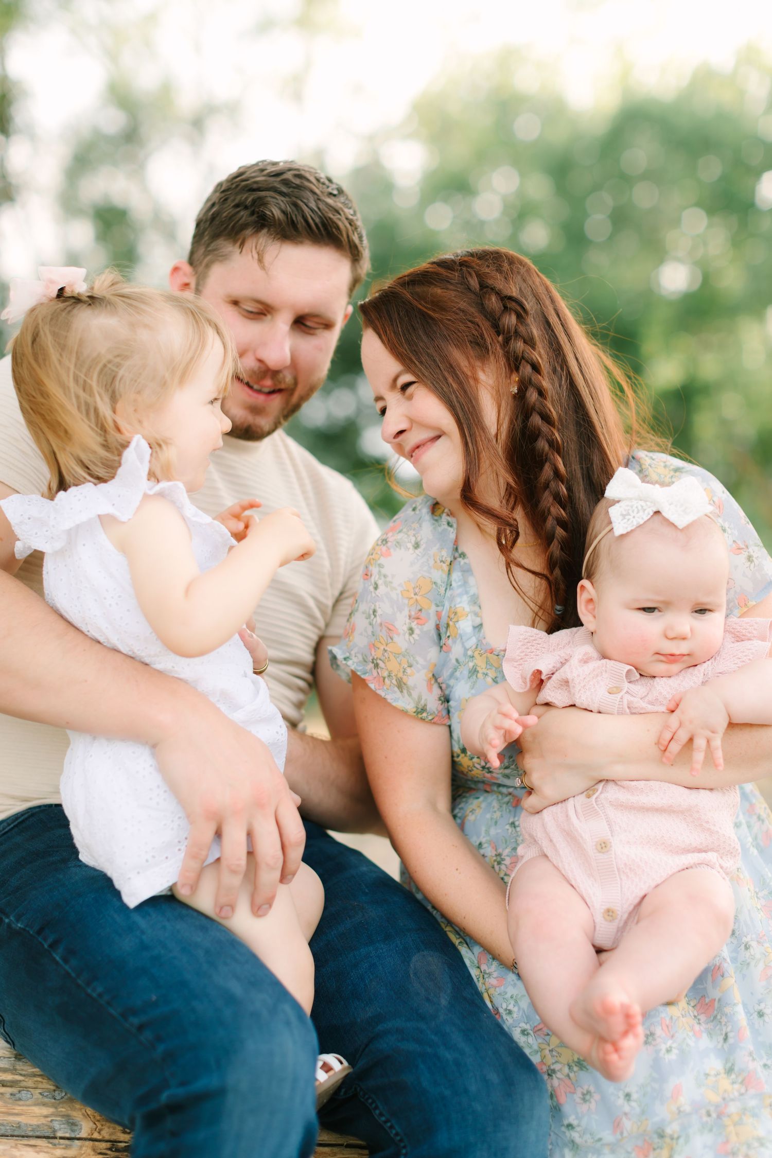 Happy young family with two baby girls sitting outdoors in soft natural light, sharing a joyful moment together.