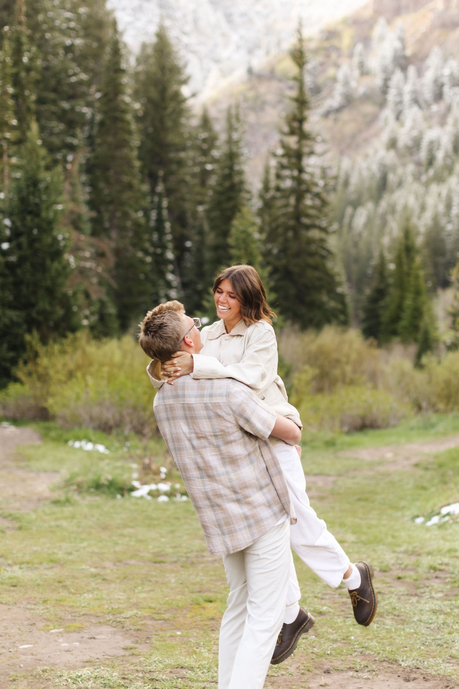 A joyful couple embraces outdoors, man lifting smiling woman against a scenic mountain forest backdrop.