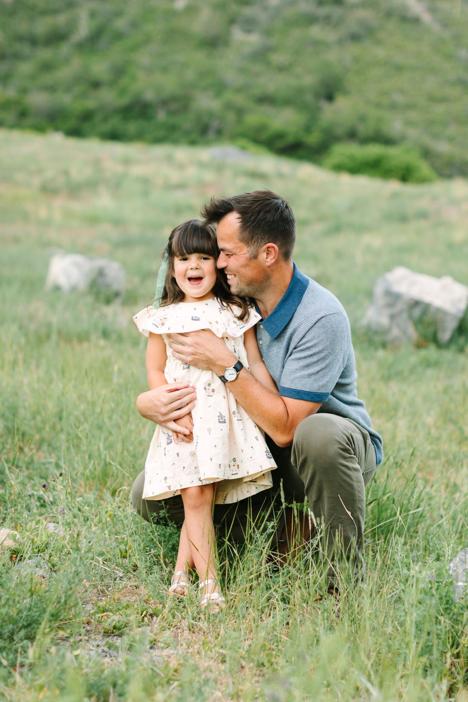 A father crouches in a green meadow, hugging his laughing young daughter in a white dress.