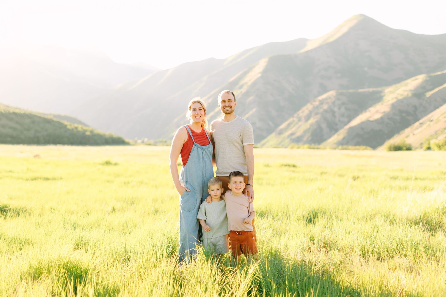 Family of four posing in a sunlit green meadow with scenic mountain backdrop during golden hour.