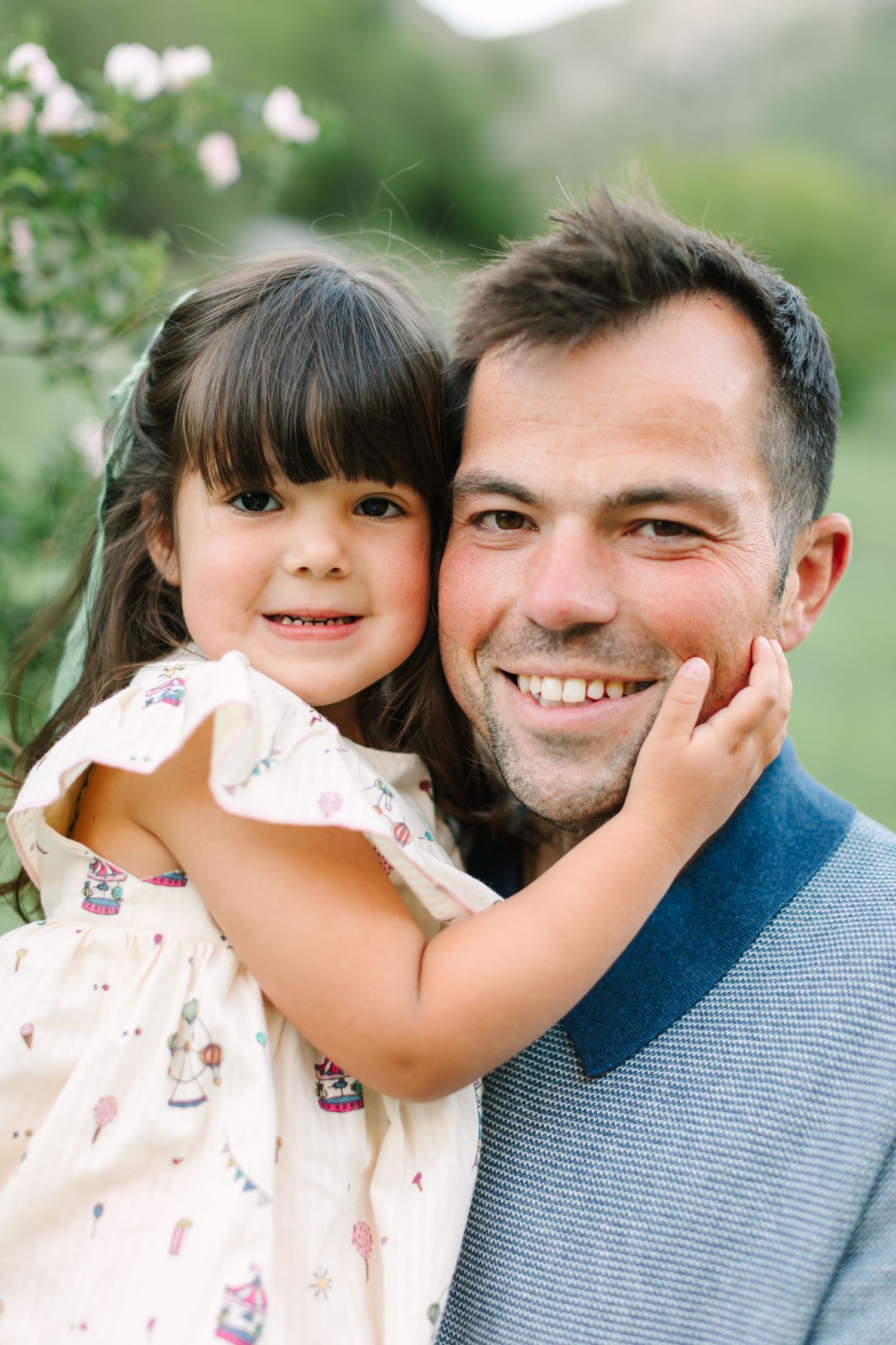 A smiling father holds his young daughter in a floral dress outdoors, as she gently cups his face with her hand.
