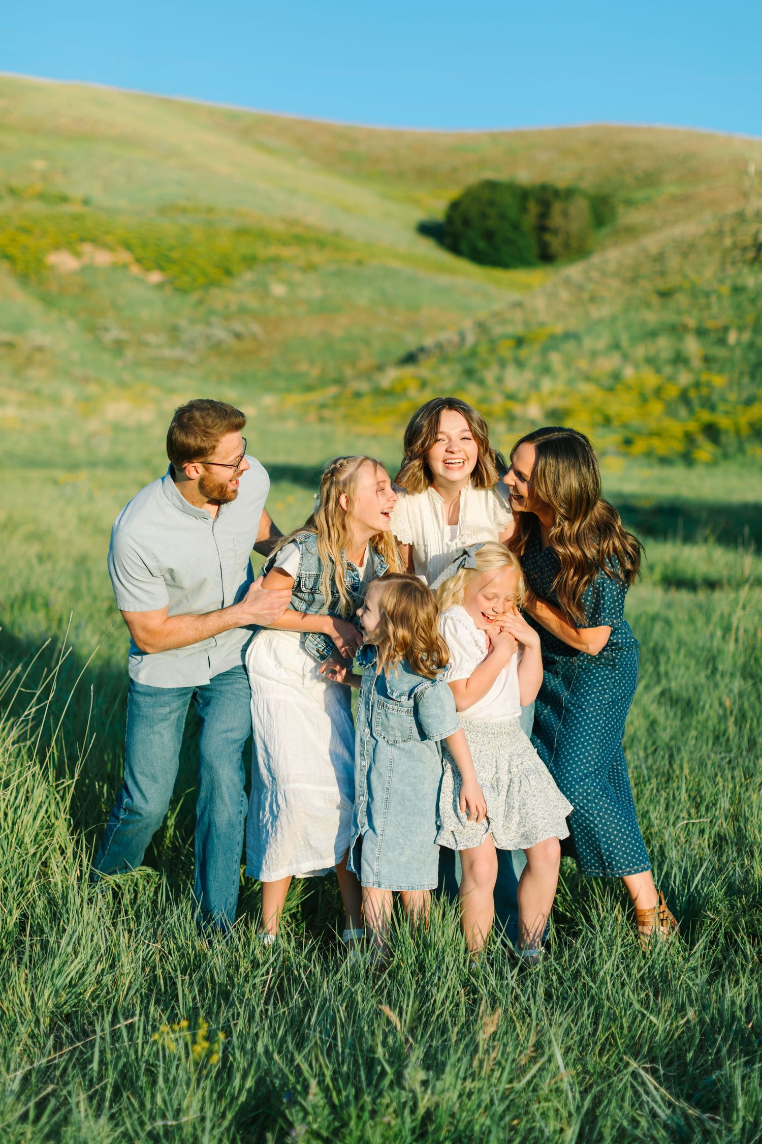 A joyful family of six laughing together in a sunny green meadow with rolling hills in the background.
