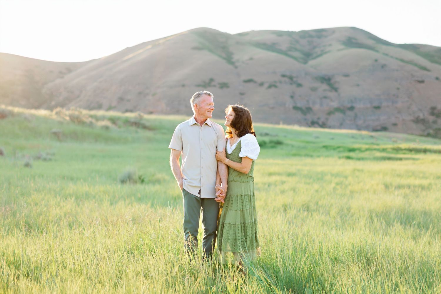 A couple stands together in a green meadow with rolling hills during golden hour sunset.