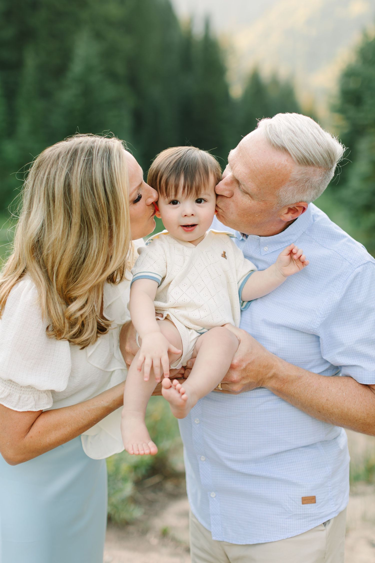 Grandparents kissing their smiling grandson on the cheeks outdoors with pine trees in the background in Utah.