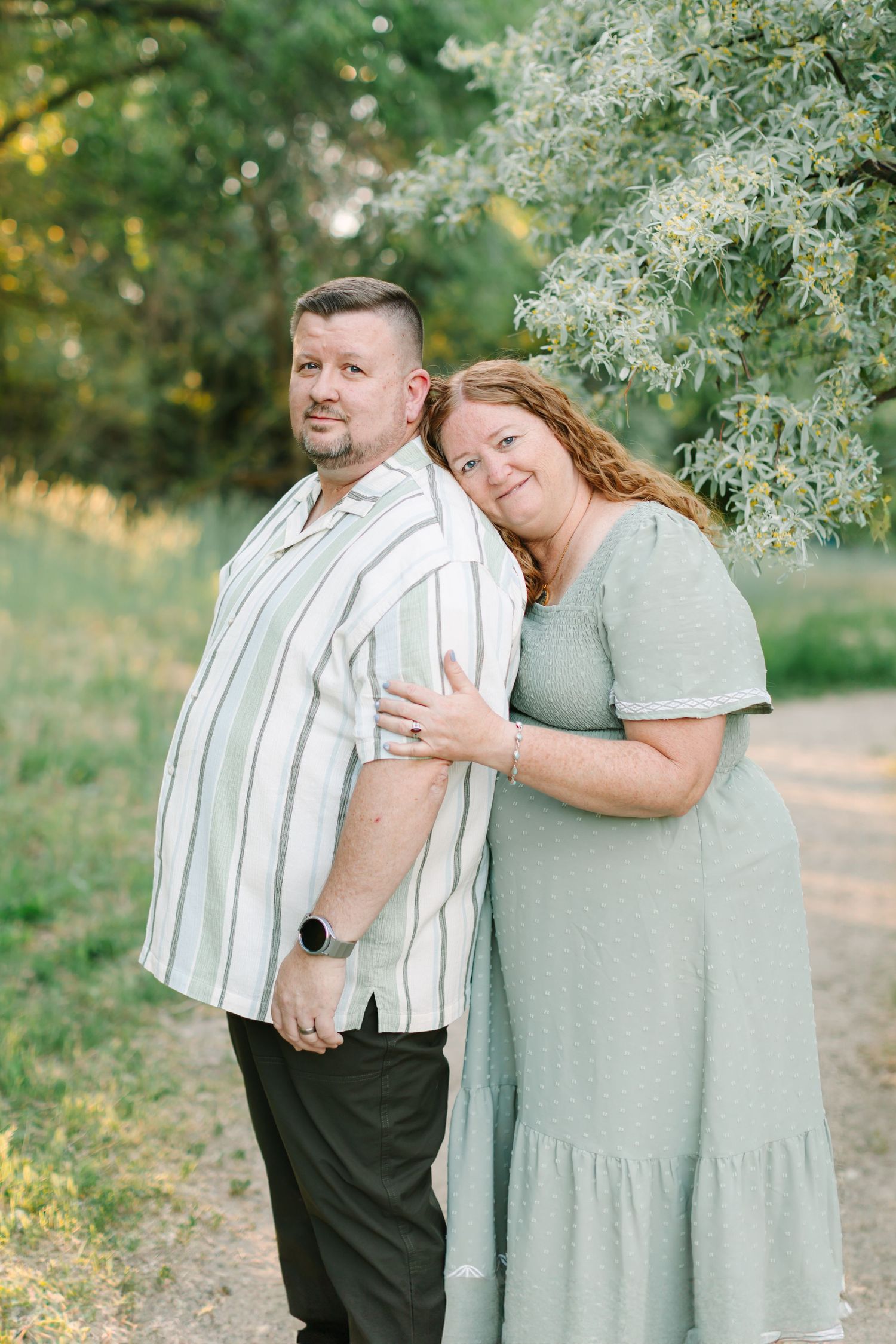 Couple embracing outdoors under green trees during a spring photo session at Wheeler Farm in Murray, Utah in Utah County photoshoot.