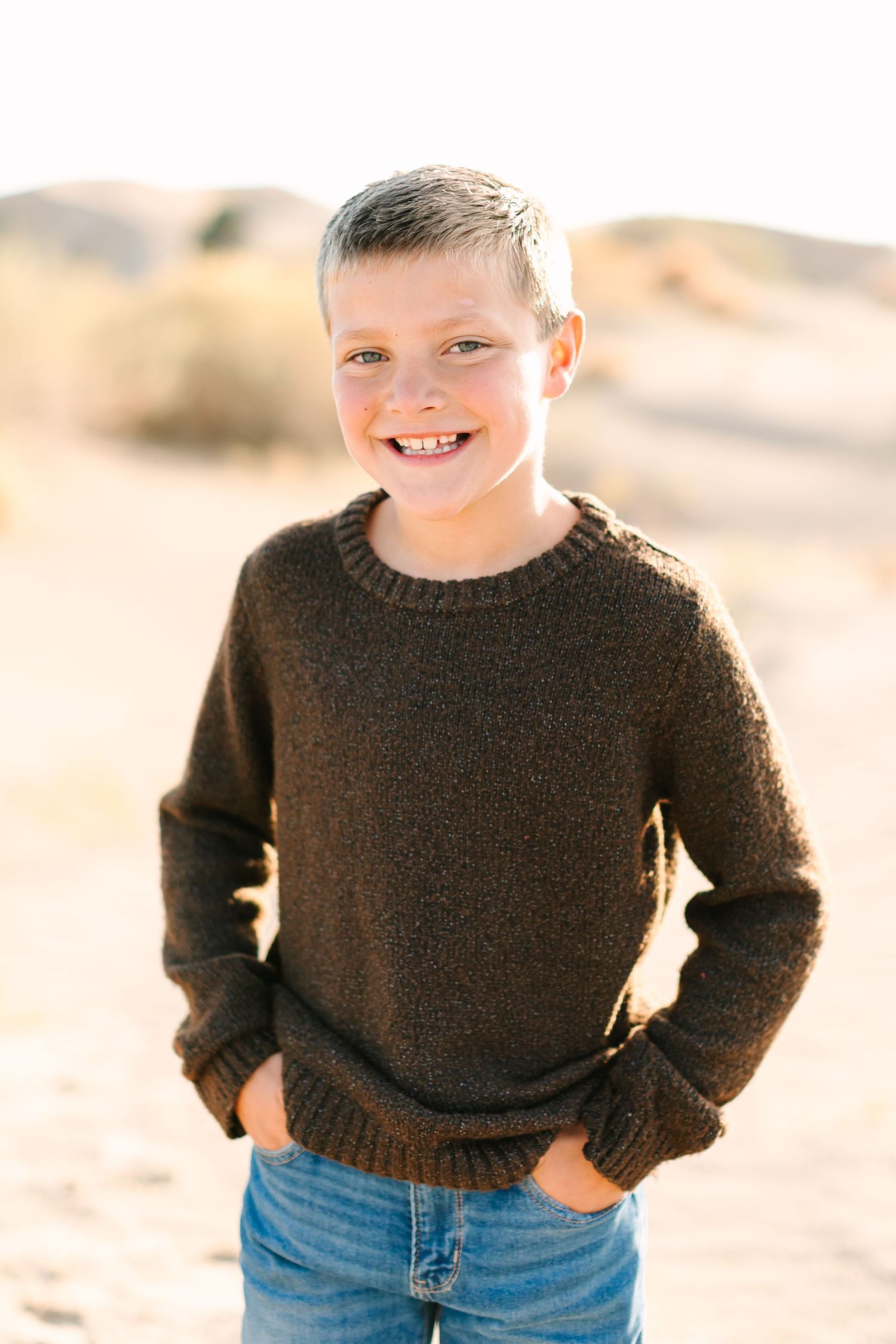 A young child wearing a brown knit sweater and blue jeans stands outdoors in natural lighting.
