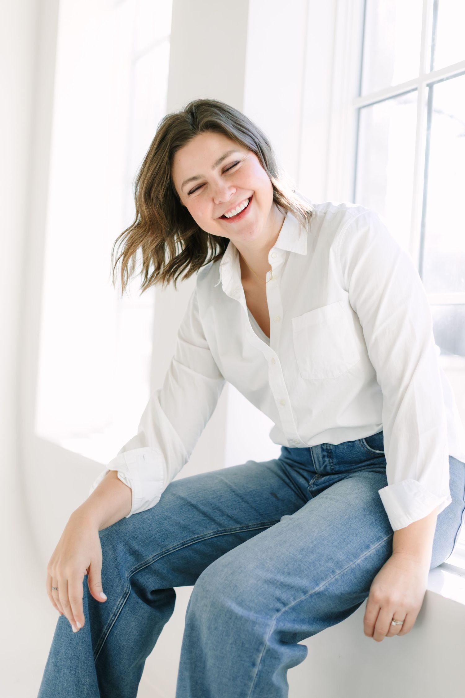 A person in a white shirt and jeans sits casually on a window ledge while smiling and looking down.