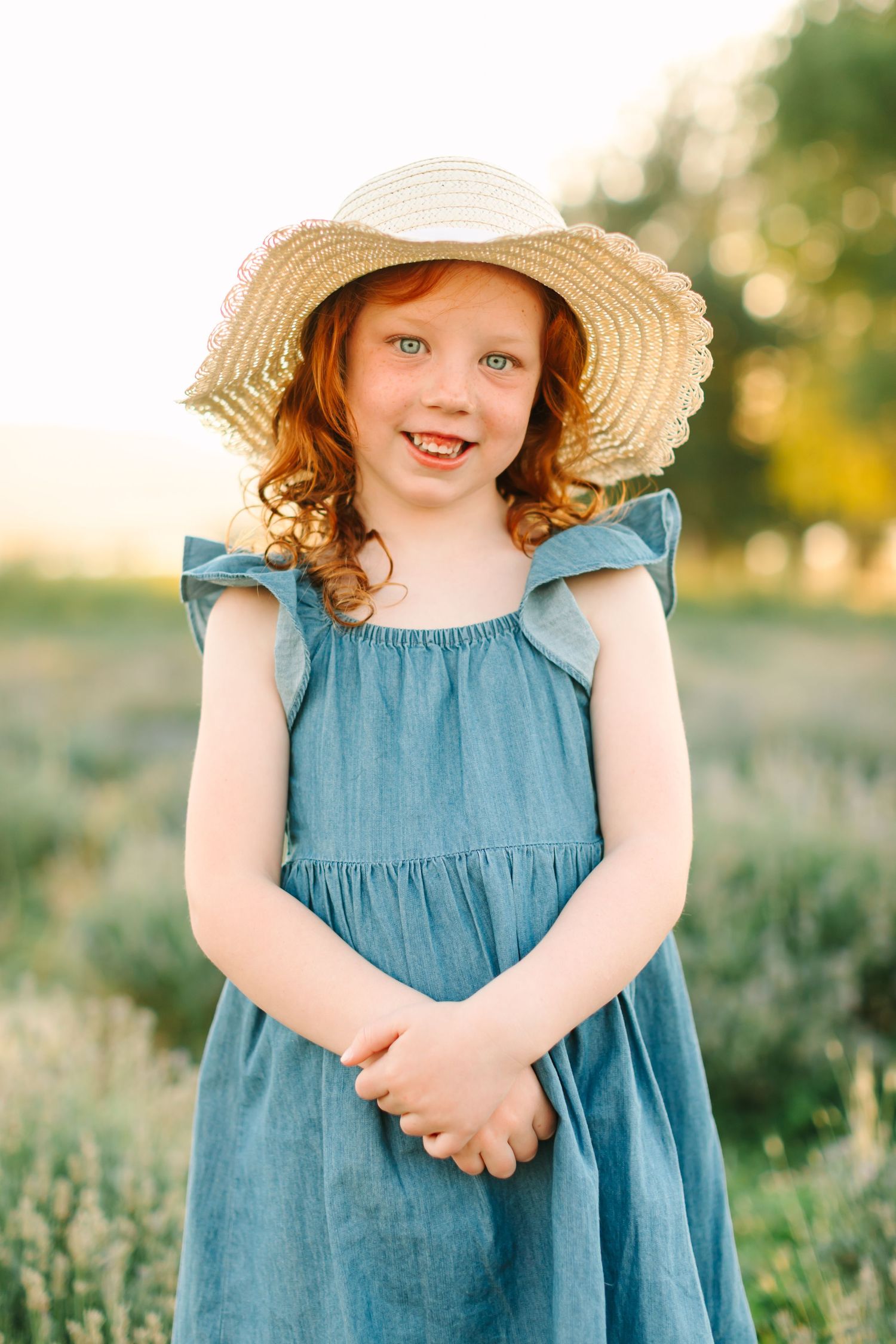 Child wearing a blue dress and straw hat stands in a sunlit meadow during golden hour.