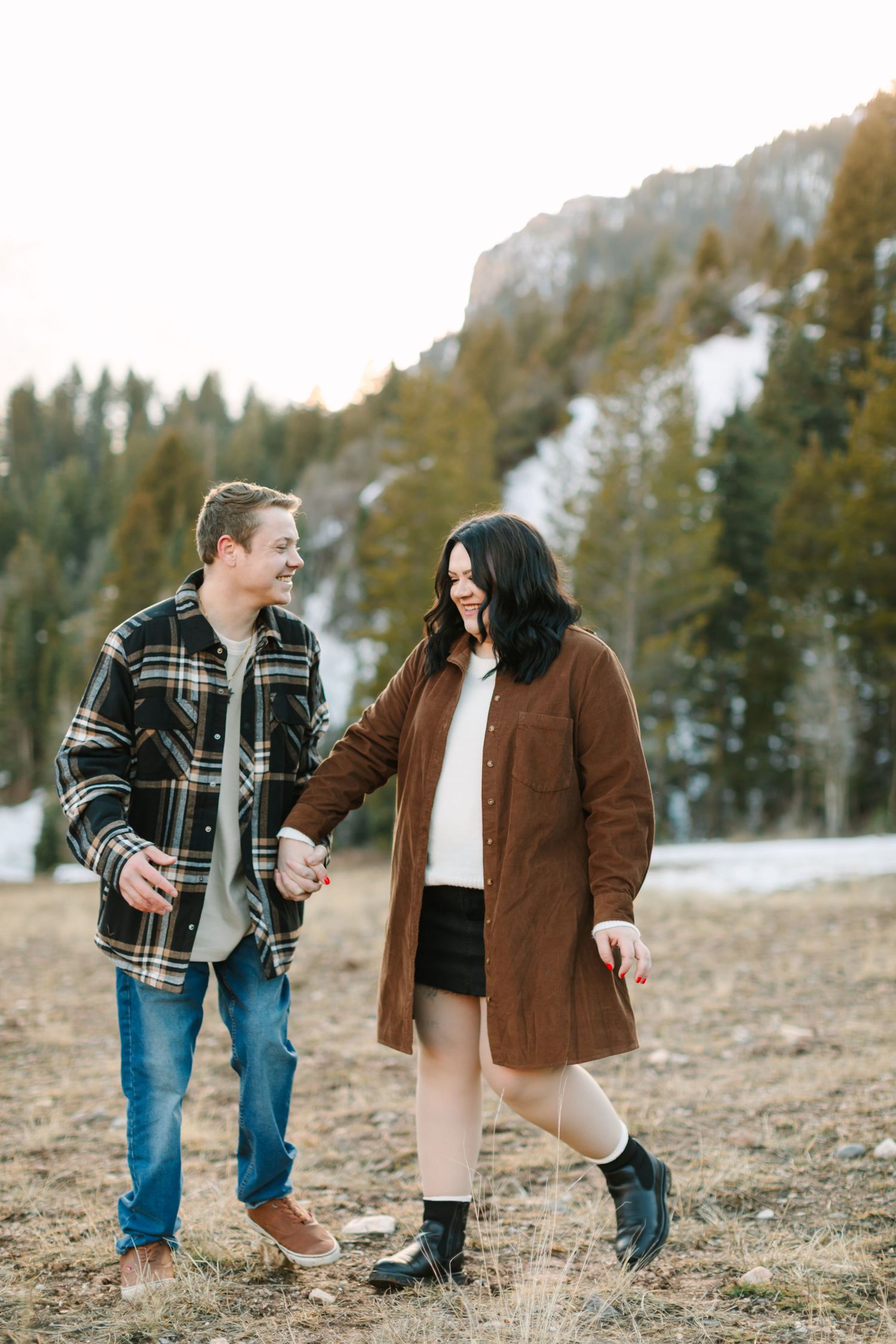 A couple in winter clothing walks together on a mountain trail with snow-capped peaks and evergreen trees in background.
