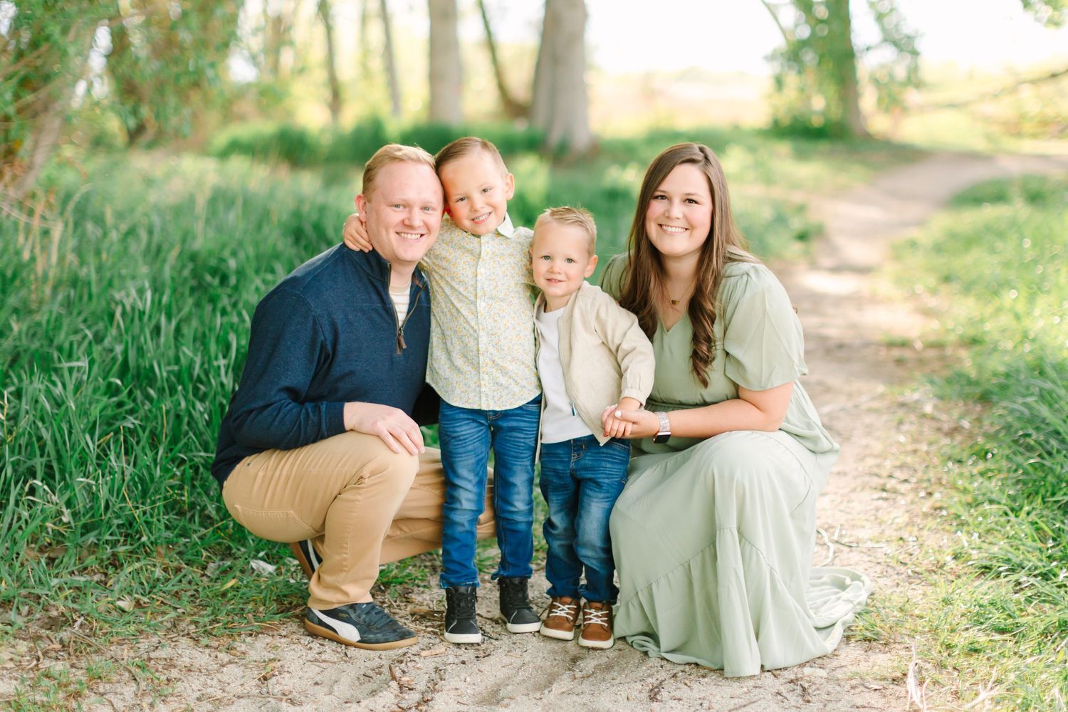A family sits together on a sunlit dirt path surrounded by tall grass and trees.