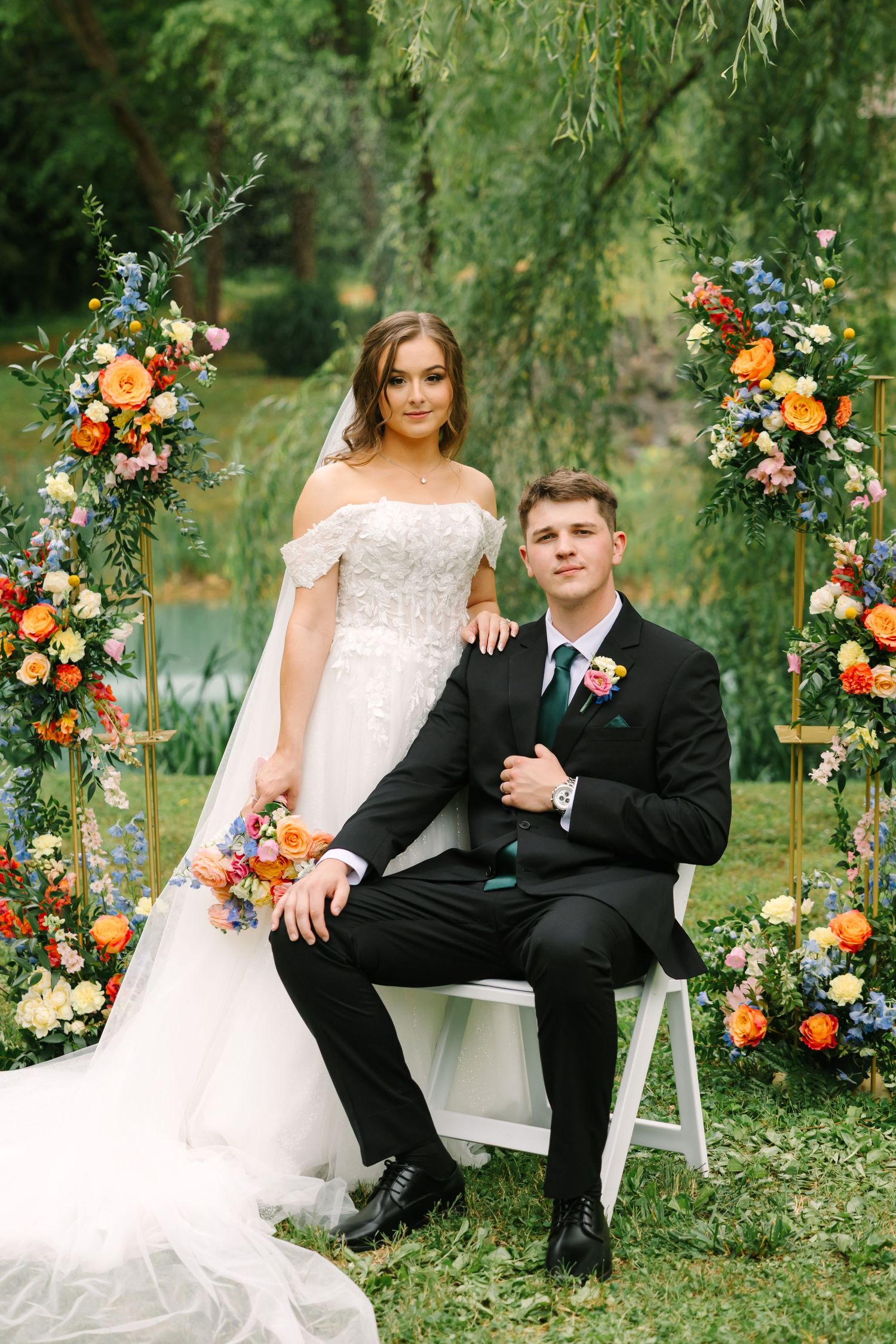 A bride and groom pose together on a white bench surrounded by floral arrangements with orange and pink blooms.