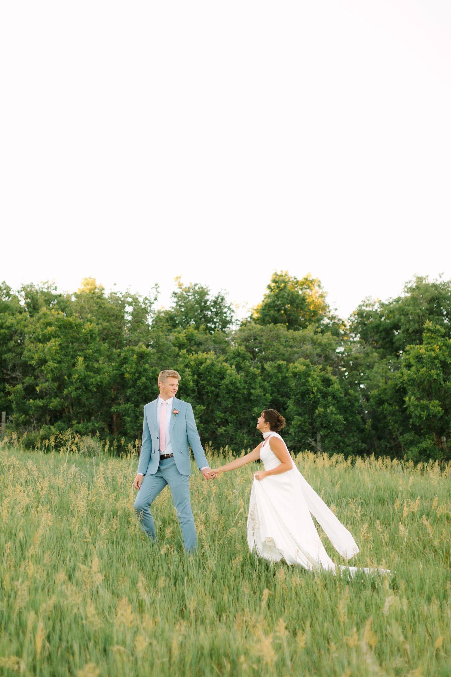 Bride and groom walk hand-in-hand through a lush green field, with the bride's white gown flowing in the breeze.