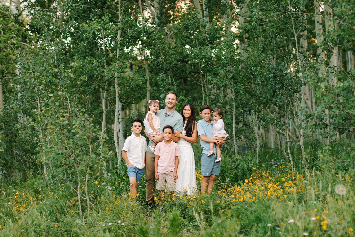 Family of seven posing outdoors among aspen trees and wildflowers in a lush green meadow in Heber, Utah.