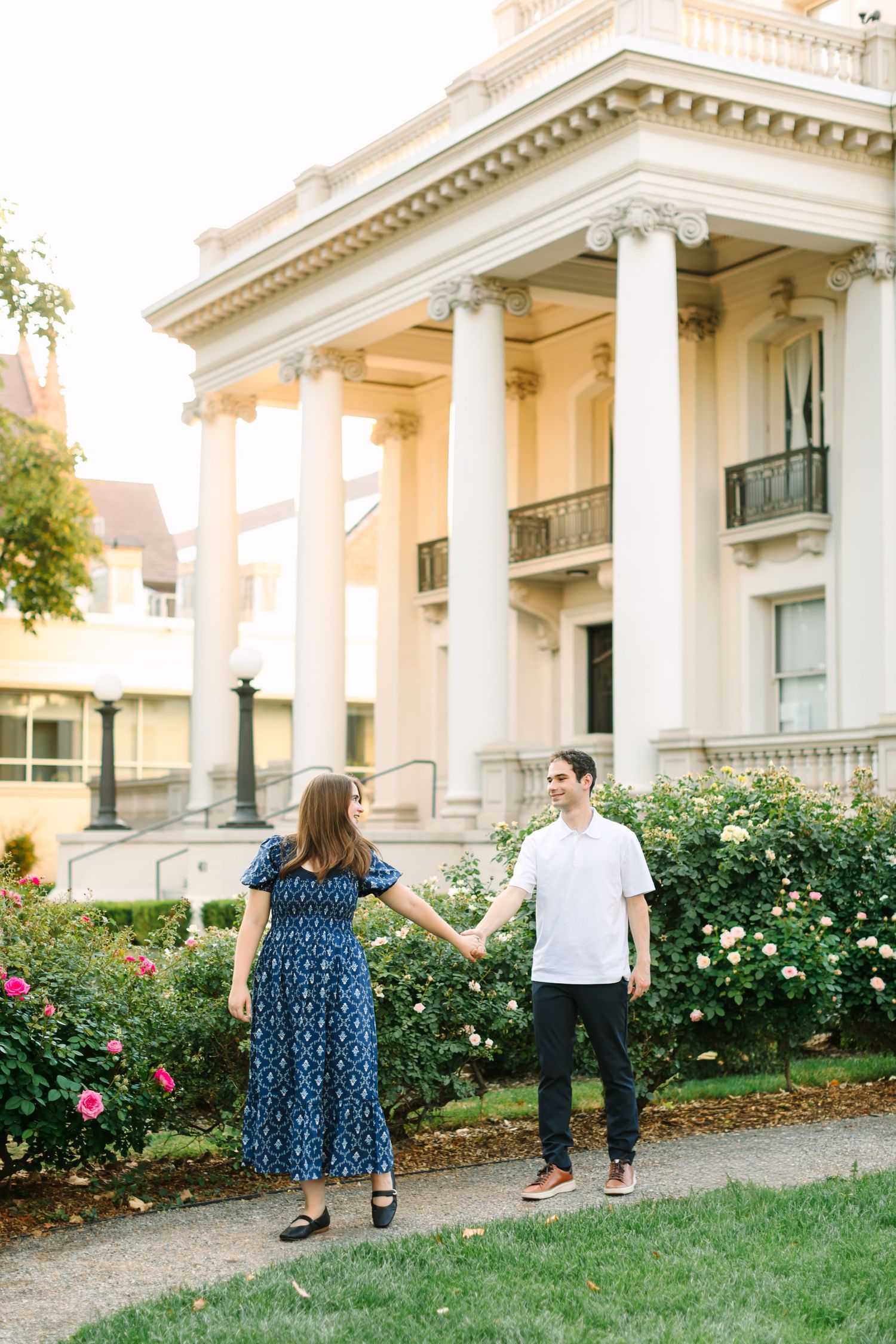 Couple holding hands smiling at each other in front of a grand white neoclassical building with columns and rose bushes in Utah.