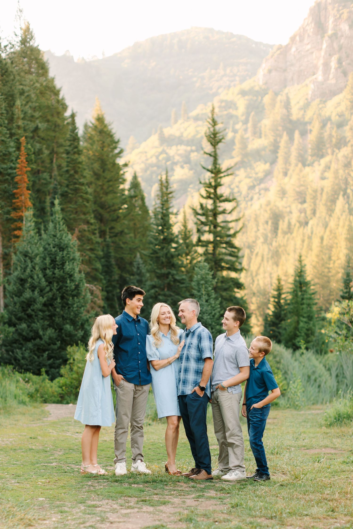 Family of six posing outdoors in mountain forest setting during golden hour, wearing coordinated blue and neutral outfits.