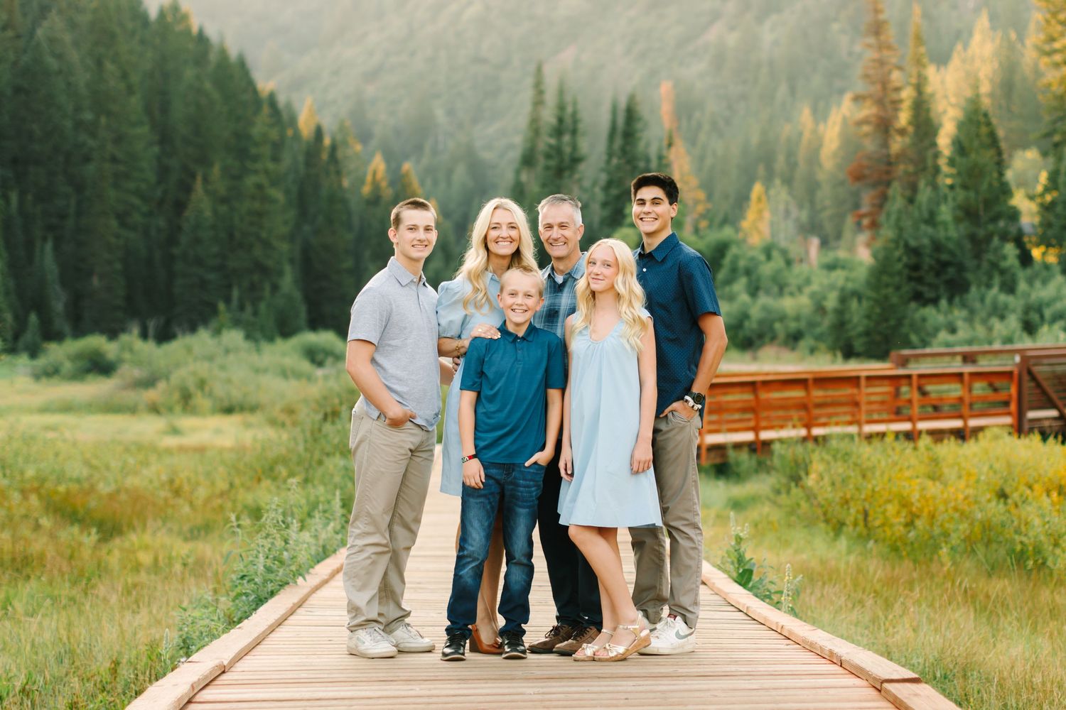 Family of six posing on a wooden boardwalk surrounded by lush green meadows and pine trees in a mountain setting.