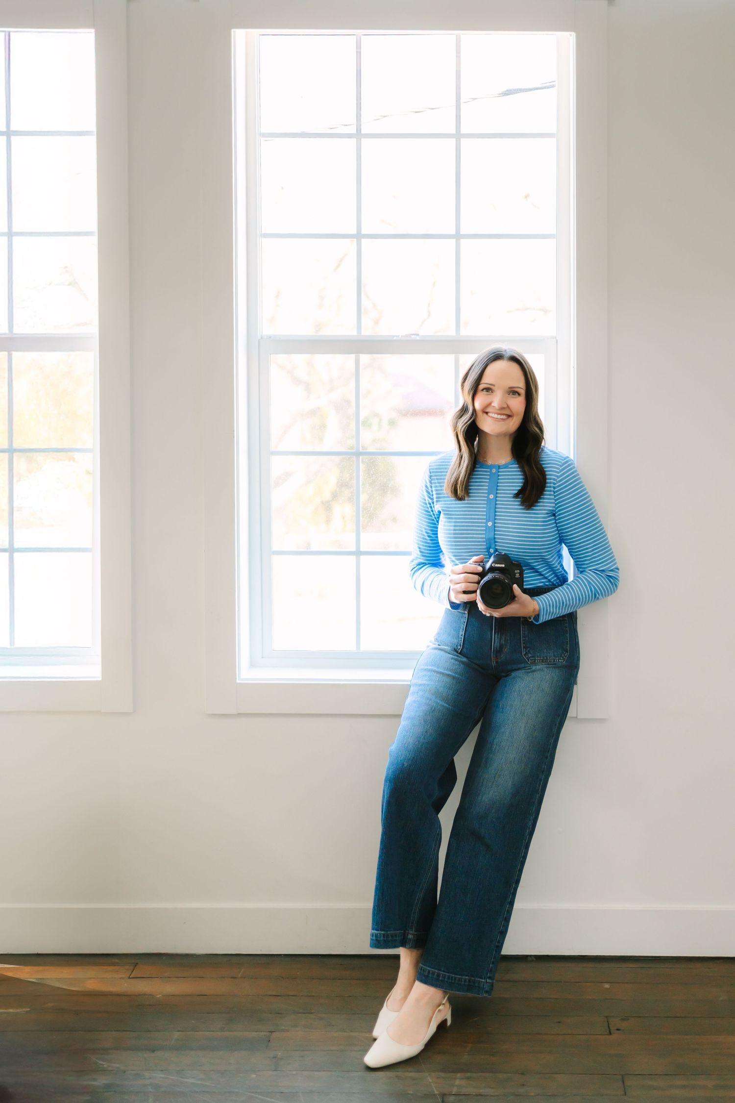A photographer in a blue sweater and wide-leg jeans stands by bright windows holding a camera in a minimalist room in Utah County
