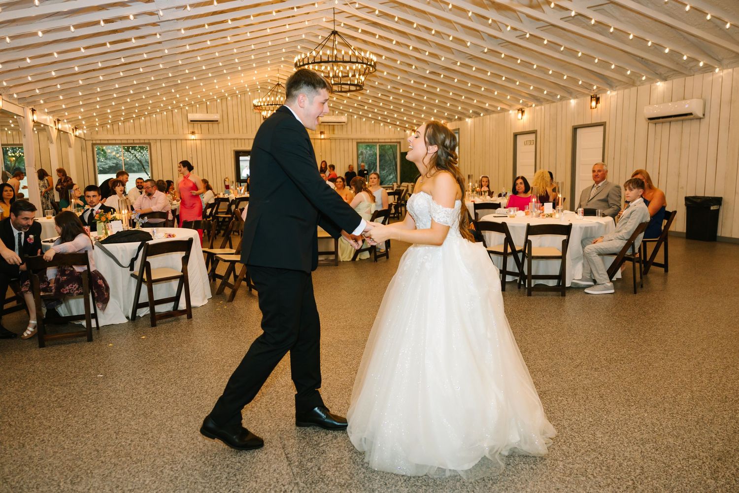 A couple performs their first dance at a wedding reception in a spacious venue with white ceiling beams and guests watching.
