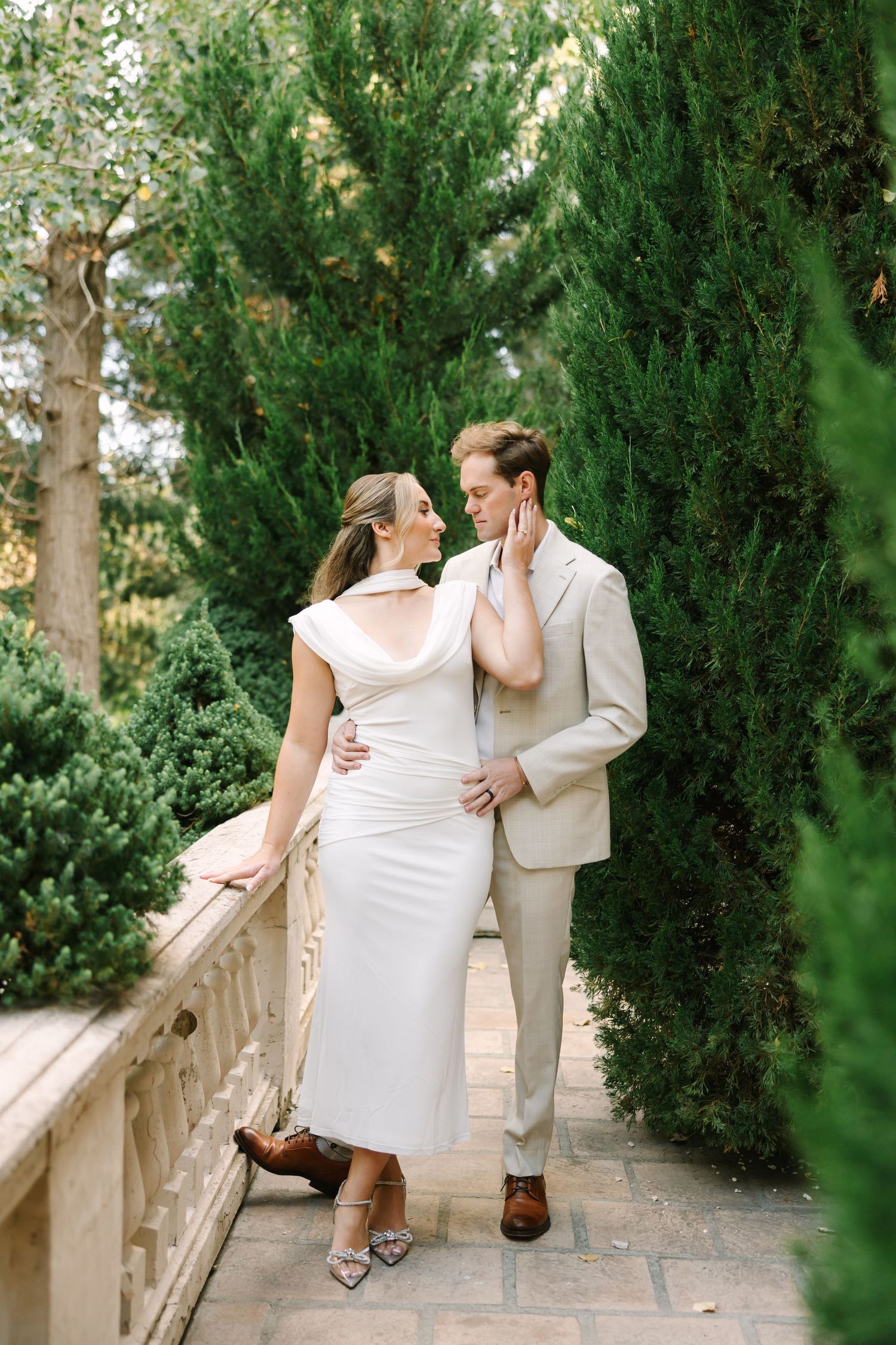 A romantic couple in light-colored formal attire embrace on a garden terrace surrounded by cypress trees.