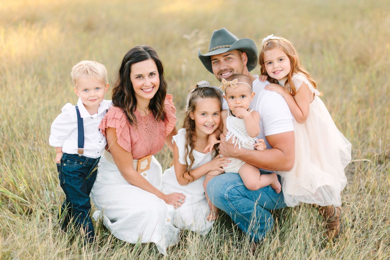 A loving family sits together in a sunlit field wearing coordinated white and blue outfits for a portrait session.