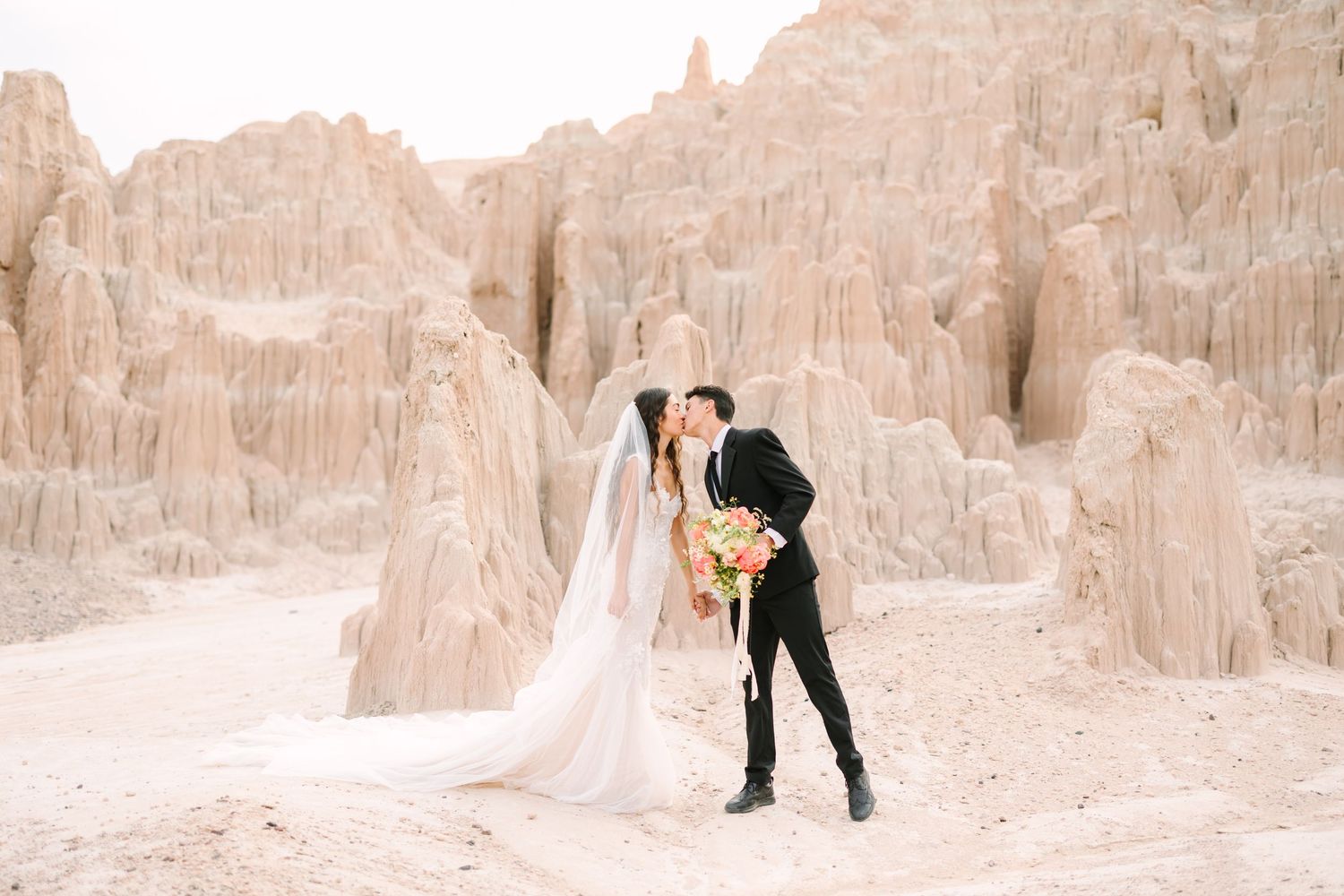 A couple in wedding attire embracing against dramatic sandstone rock formations in a desert landscape.