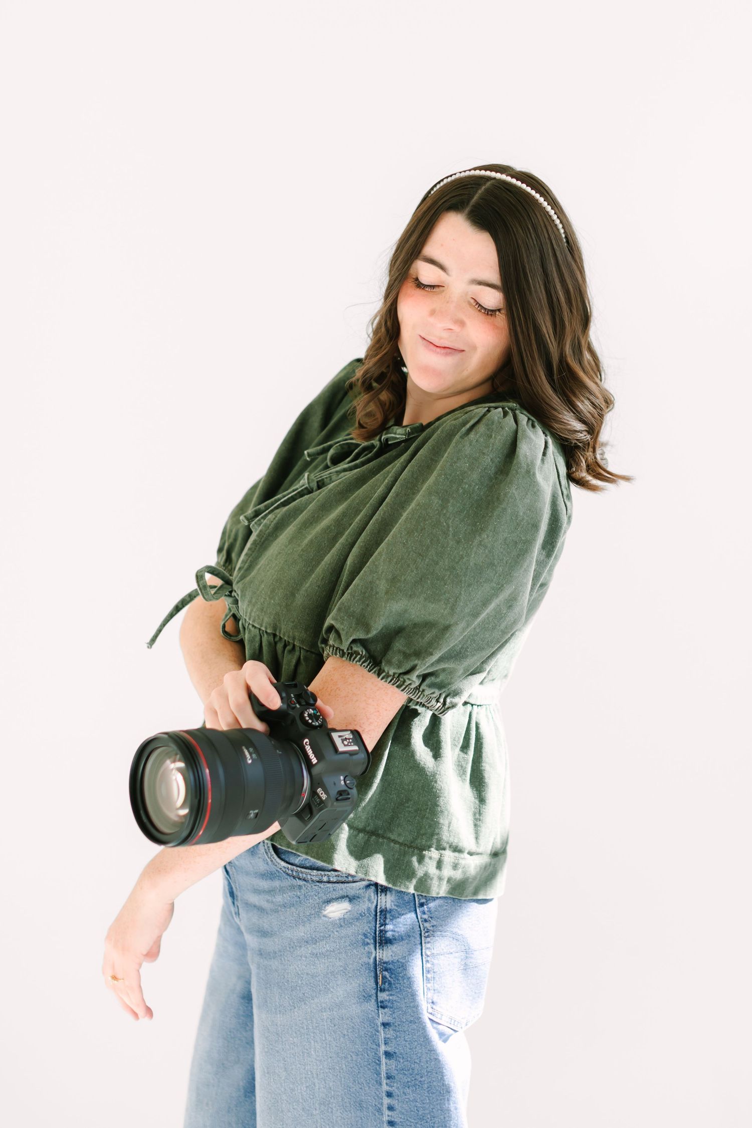 A photographer wearing a green sweater and jeans holds a professional camera while posing against a white background.