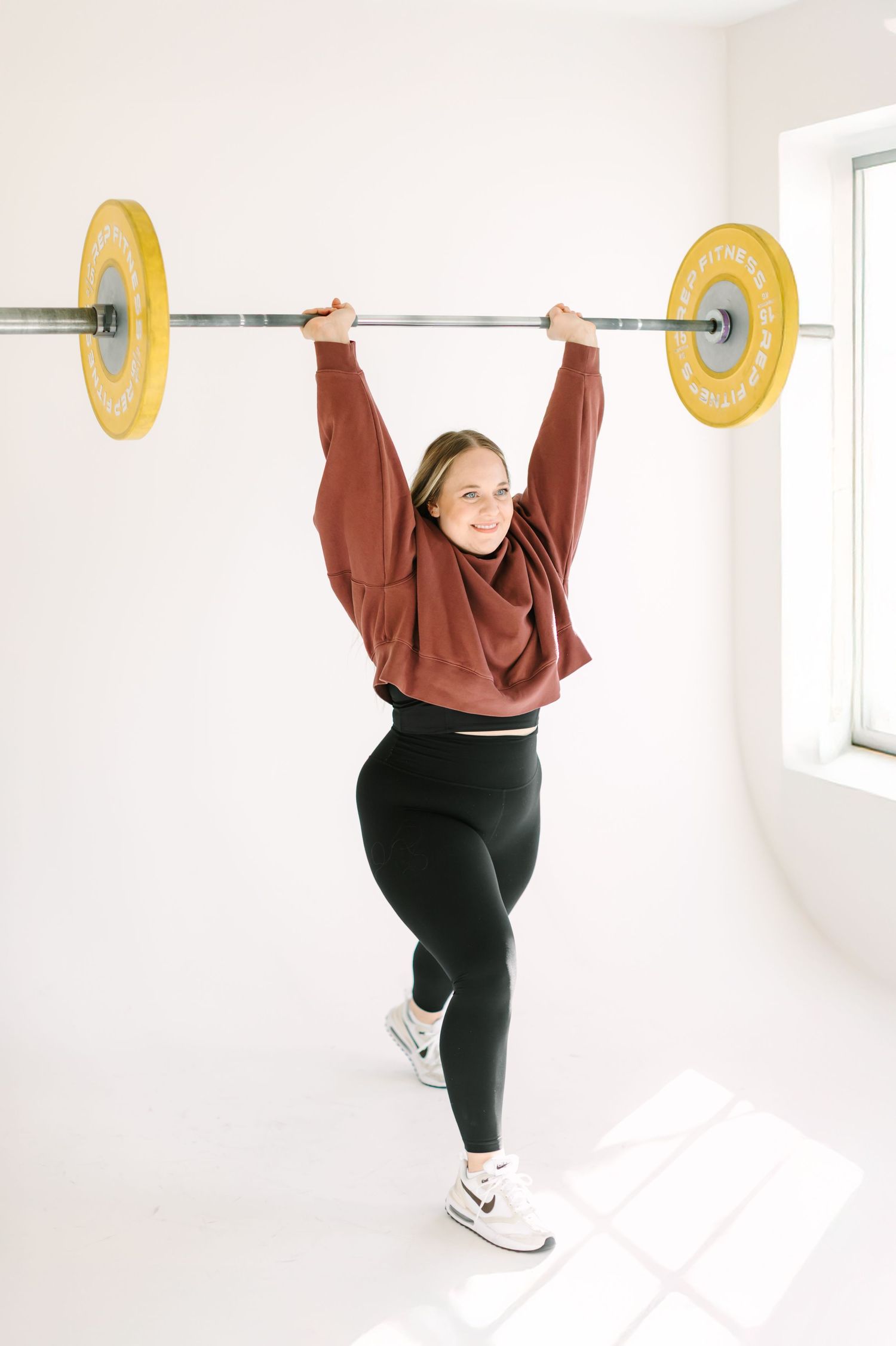 A series of four progressive images showing someone lifting a barbell overhead in a white room while wearing brown top and black pants.