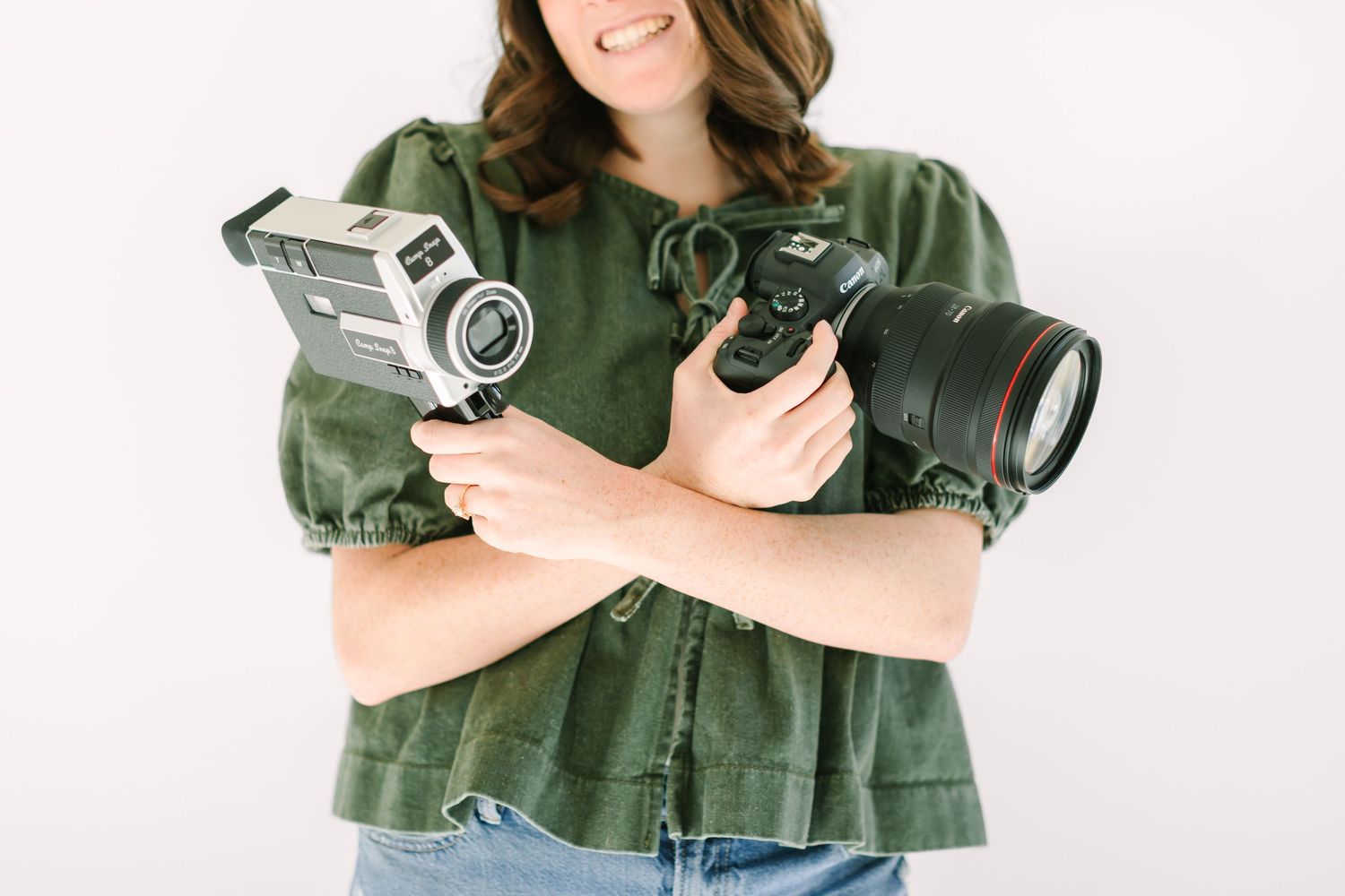 A person wearing a green top holds a professional camera while posing against a white background.