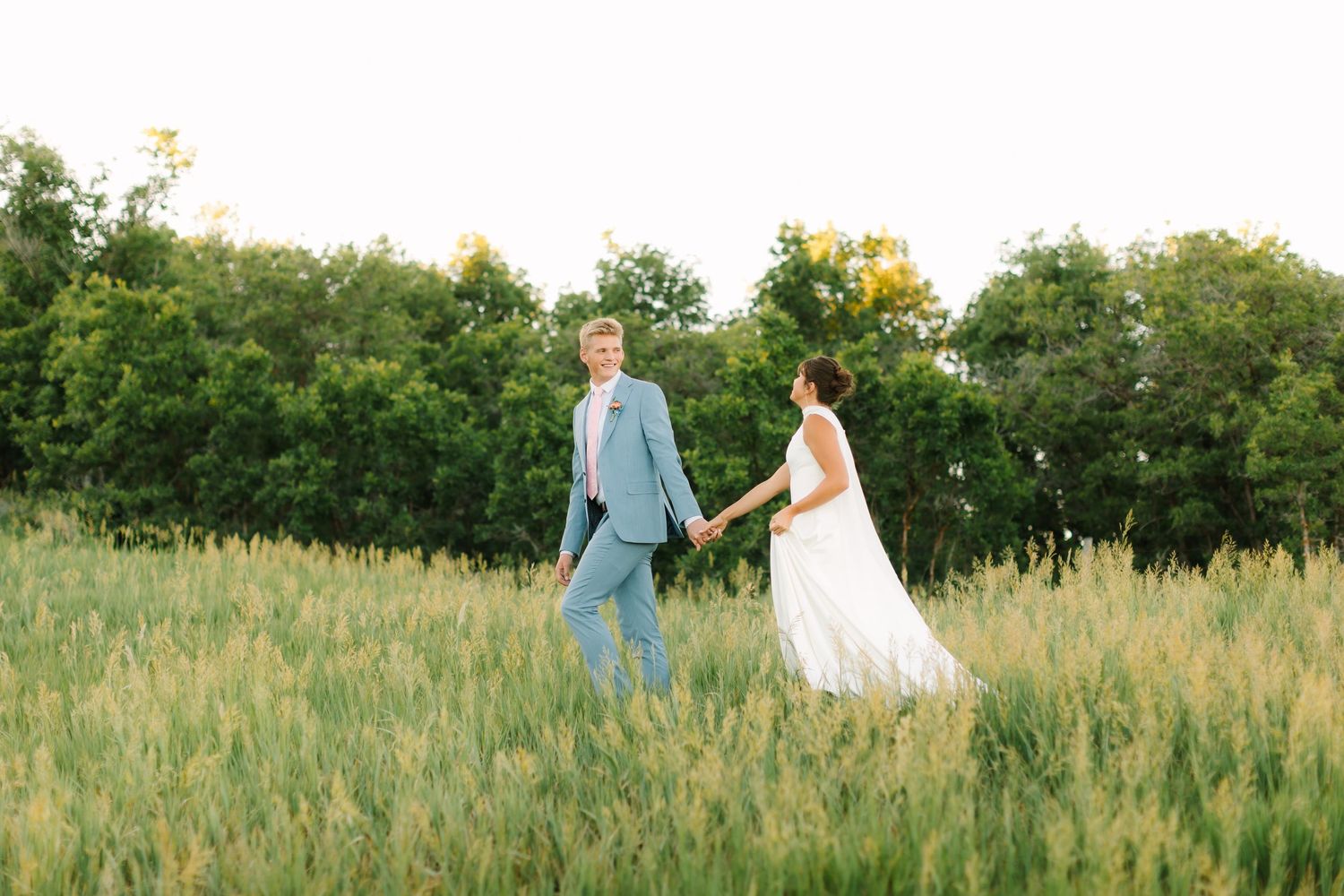 Bride and groom holding hands walking through lush green field at golden hour in wedding attire.