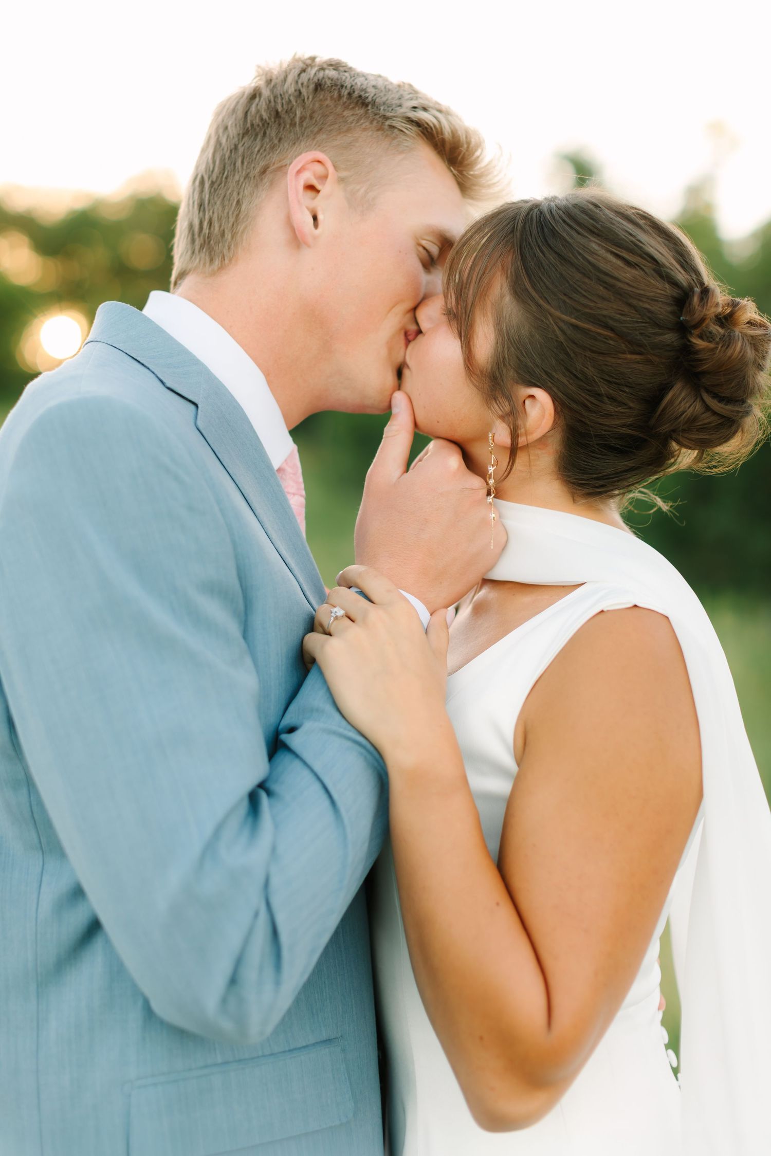 Bride and groom share a romantic kiss outdoors, with the groom in a light blue suit and bride in a white dress.