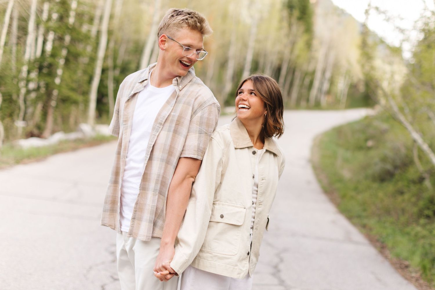 Happy couple holding hands and laughing while walking on a scenic tree-lined path outdoors.