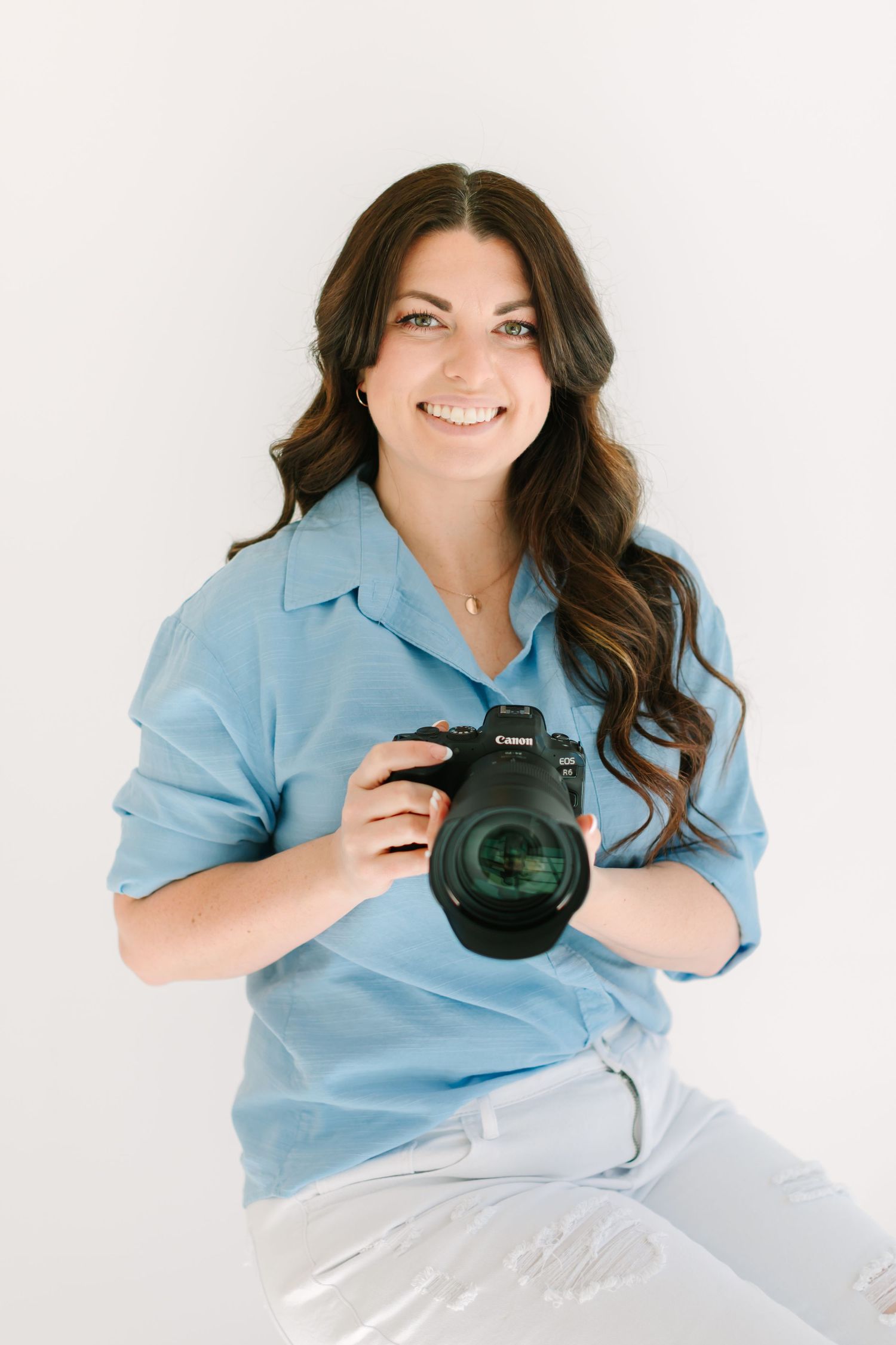 Photographer wearing blue button down shirt holds professional camera against white backdrop.
