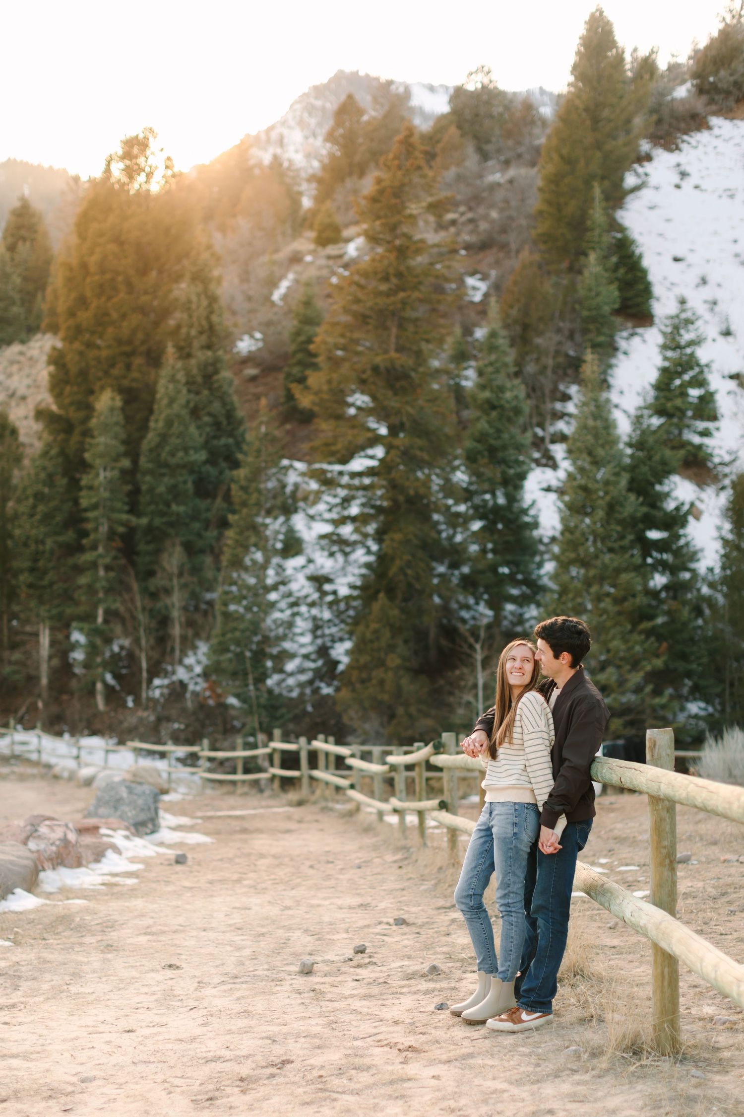 A romantic winter engagement photo session in the snowy mountains with evergreen trees and a rustic wooden fence.
