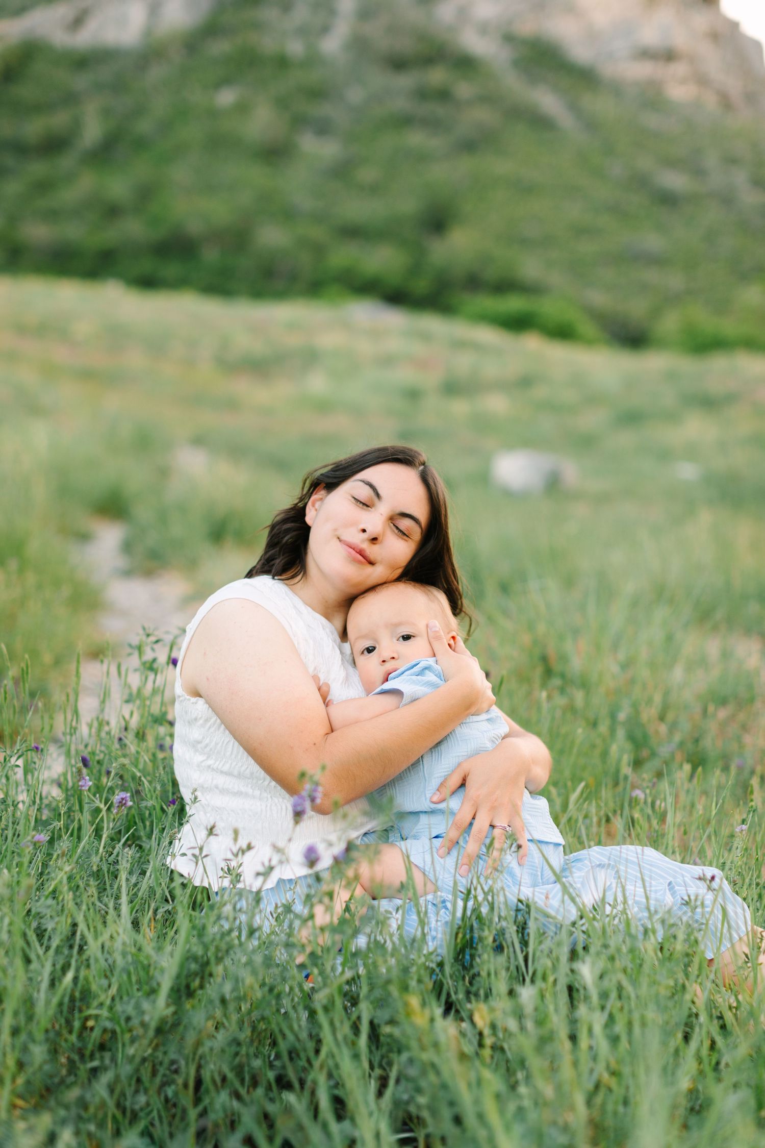 A mother with dark hair sits in a green meadow, tenderly embracing her baby in a heartwarming outdoor moment.