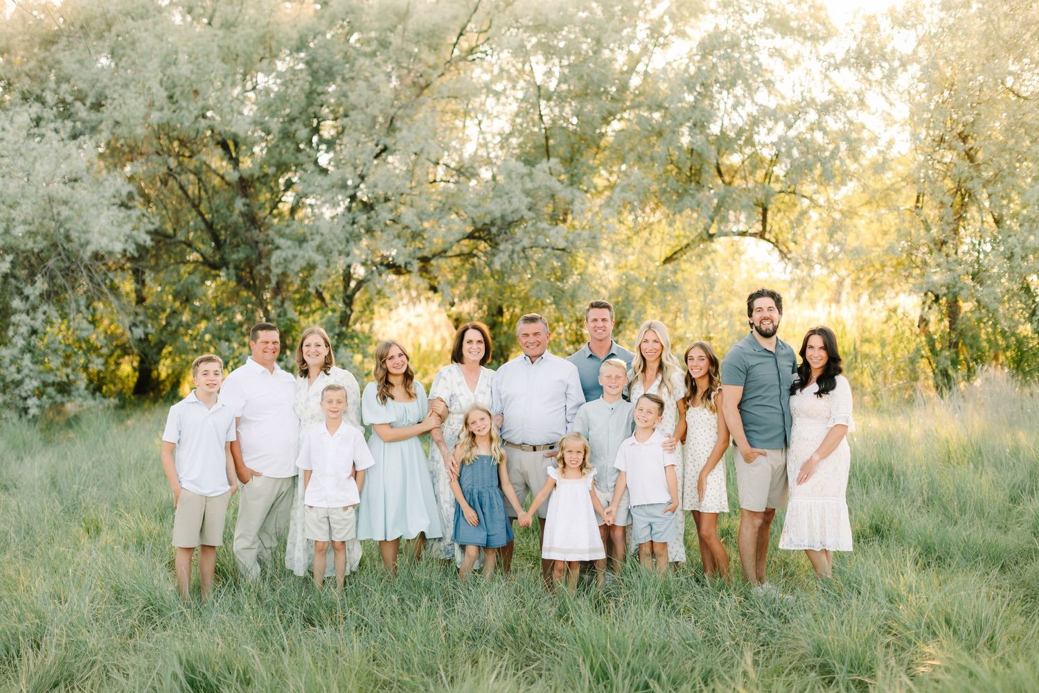 Large family portrait in a sunlit meadow with lush green trees, wearing coordinated white and pastel outfits in Bountiful, Utah.