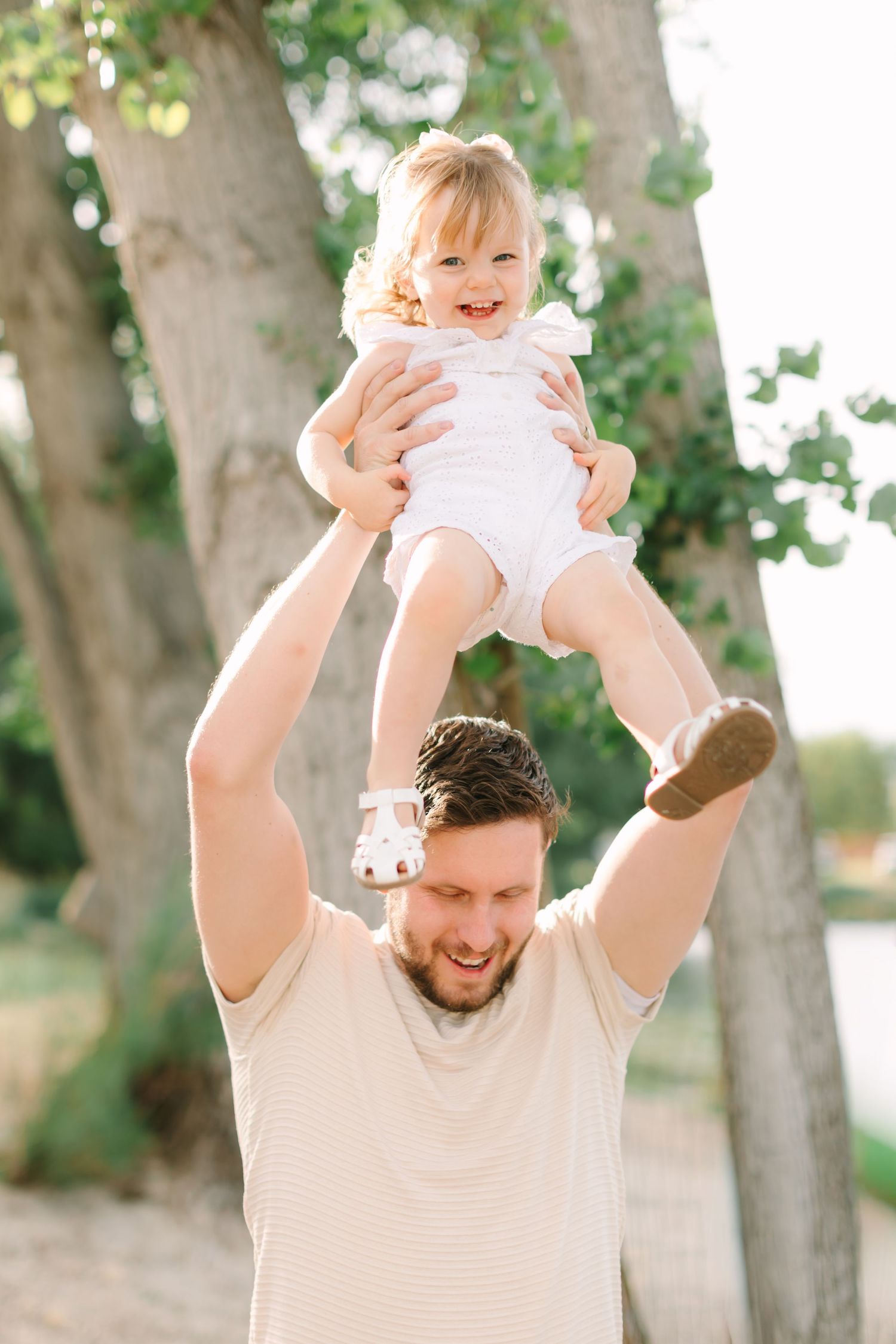 A smiling father lifts his laughing toddler daughter high above his head outdoors near trees on a sunny day.