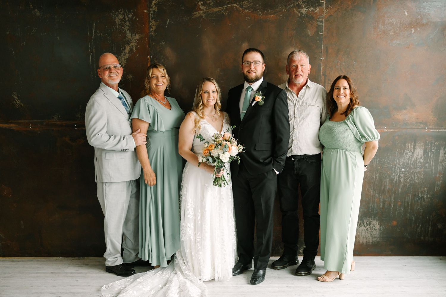 Wedding party members in sage green dresses and dark suits pose together in an industrial-style venue.