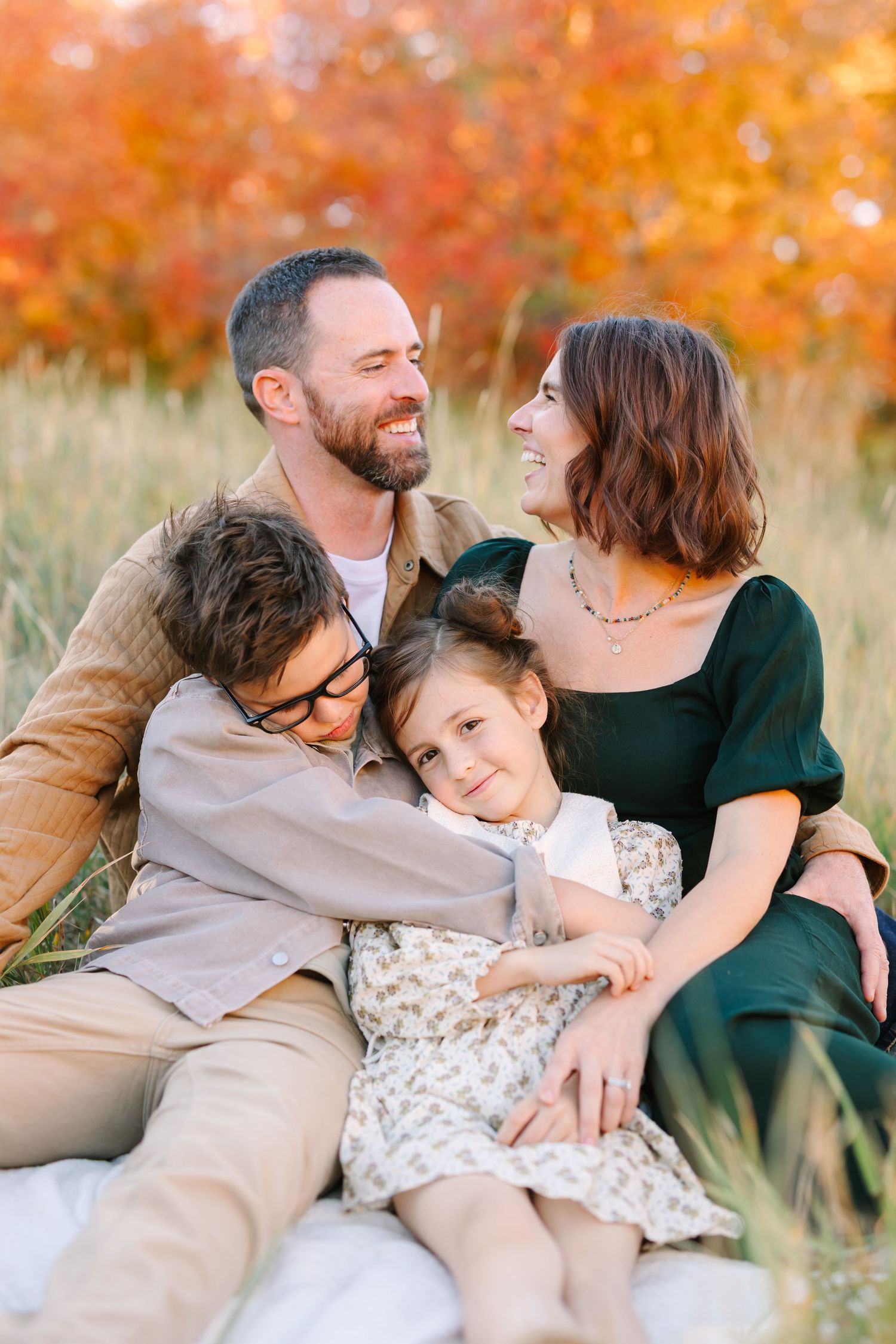 A family sits together in a field during autumn with vibrant orange foliage in the background.