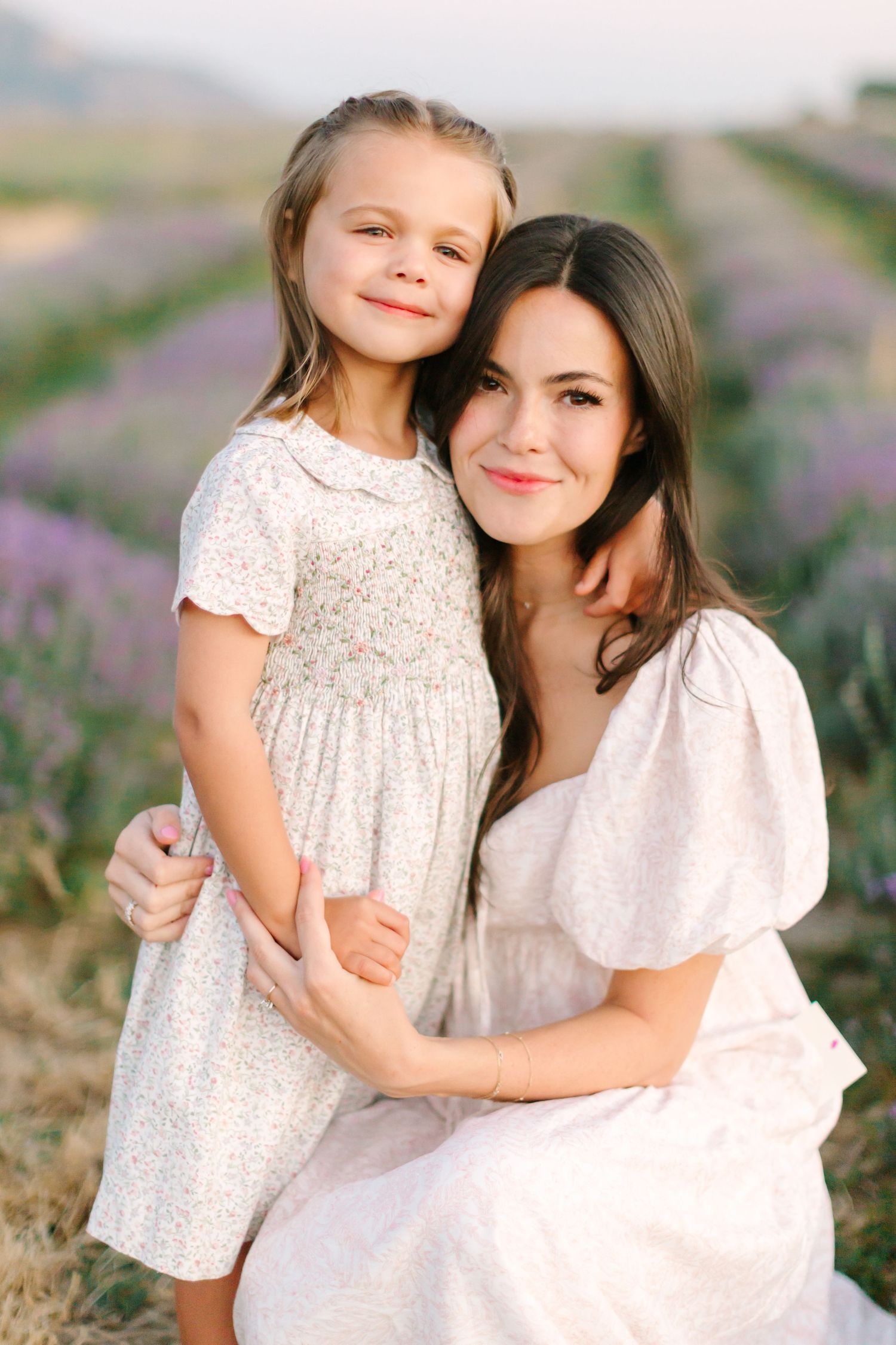 Parent and child in white dresses pose together in a blooming lavender field during sunset.
