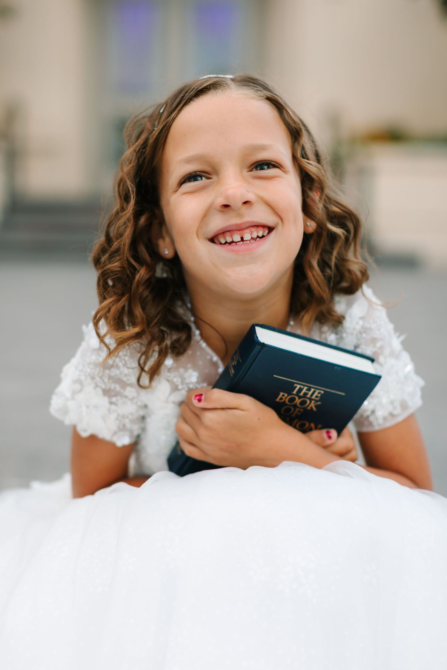 A child in white lace holds a Bible while lying on white fabric with a bright, cheerful expression.