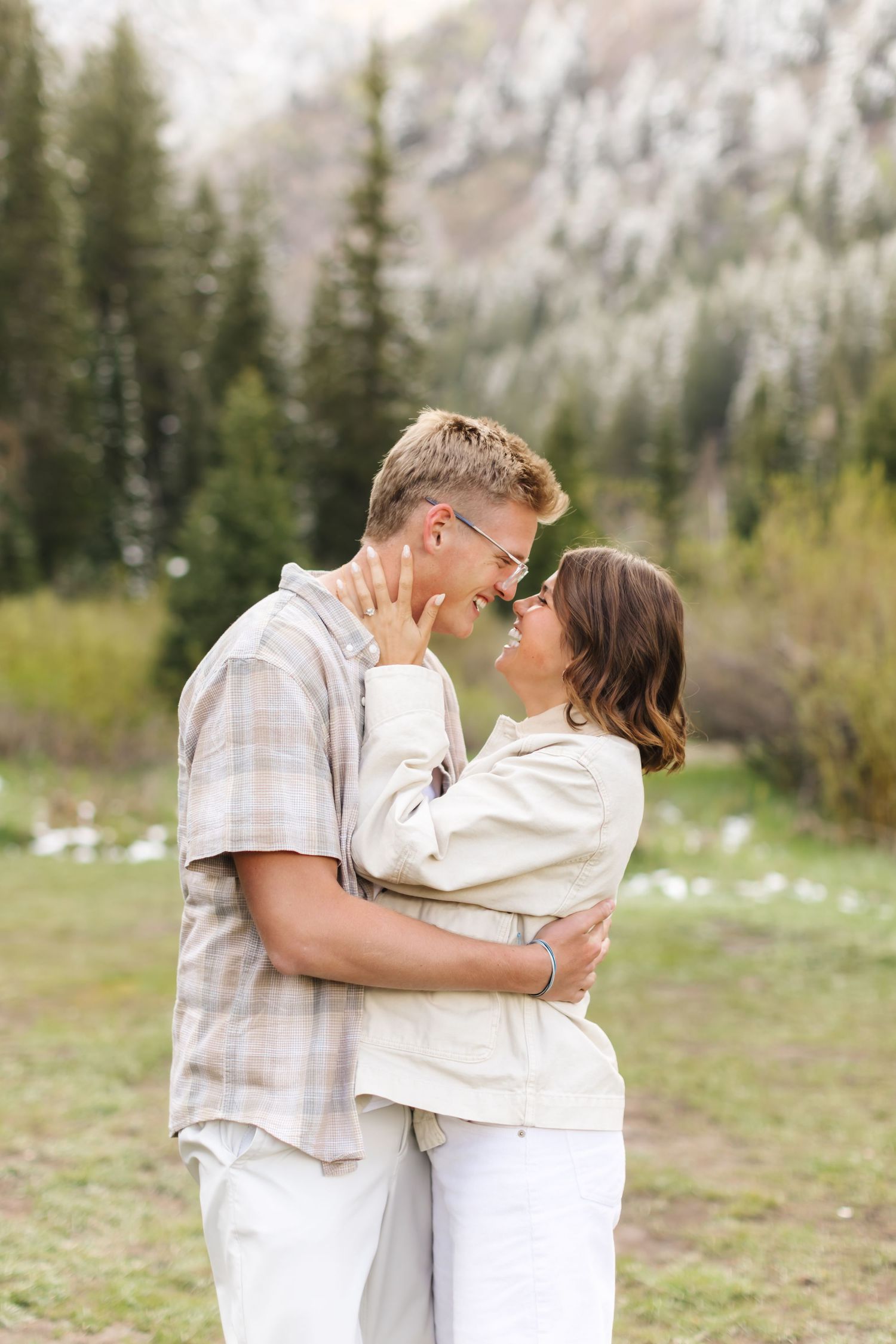Couple embracing and smiling at each other in a scenic mountain meadow with evergreen trees in the background.
