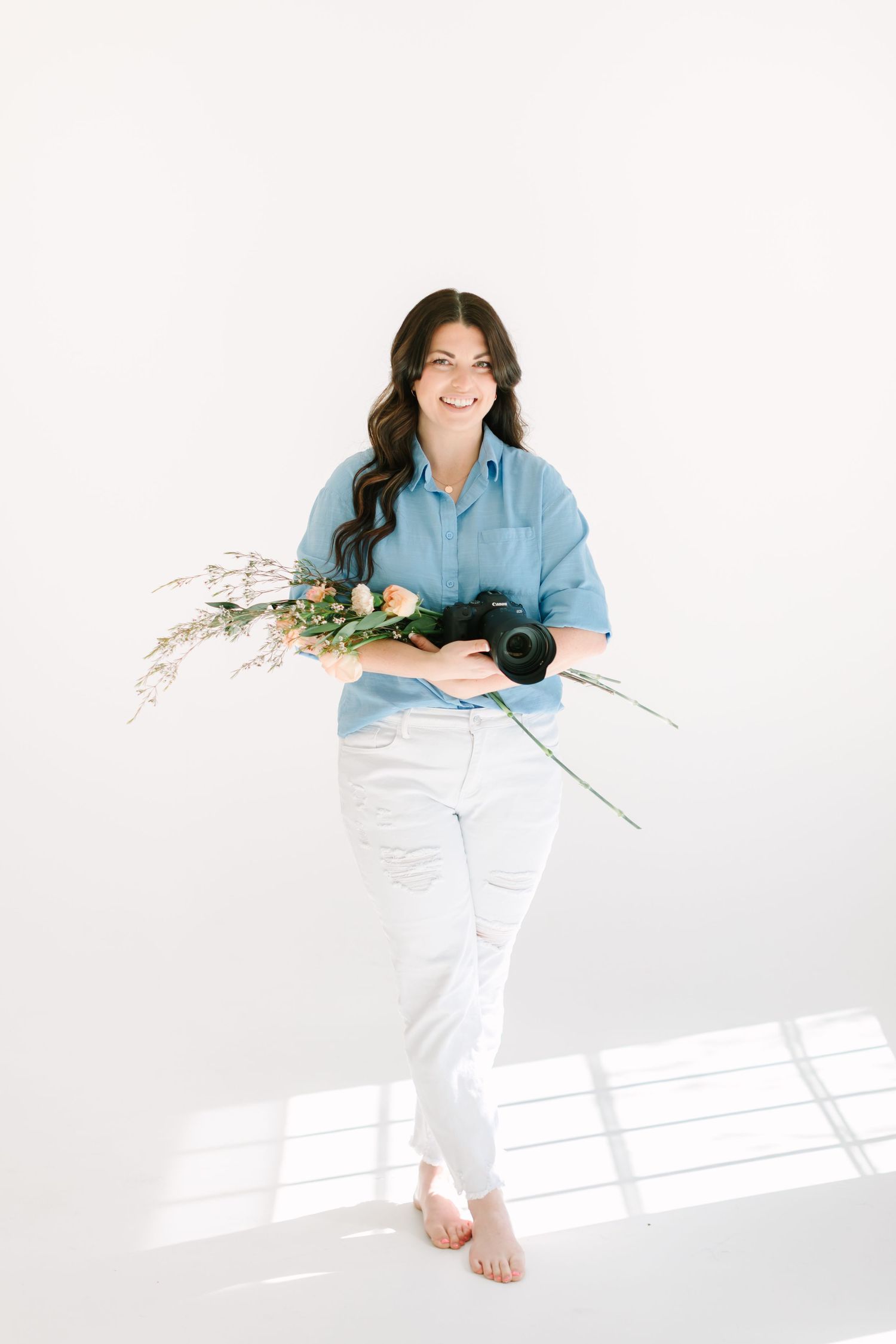 Photographer in casual attire holds camera and floral stems while sitting on white floor.