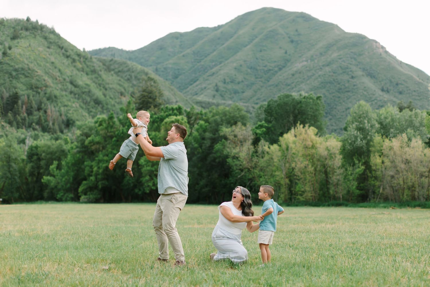 Family enjoying a candid outdoor photoshoot in a scenic mountain meadow with lush green grass and rolling hills.